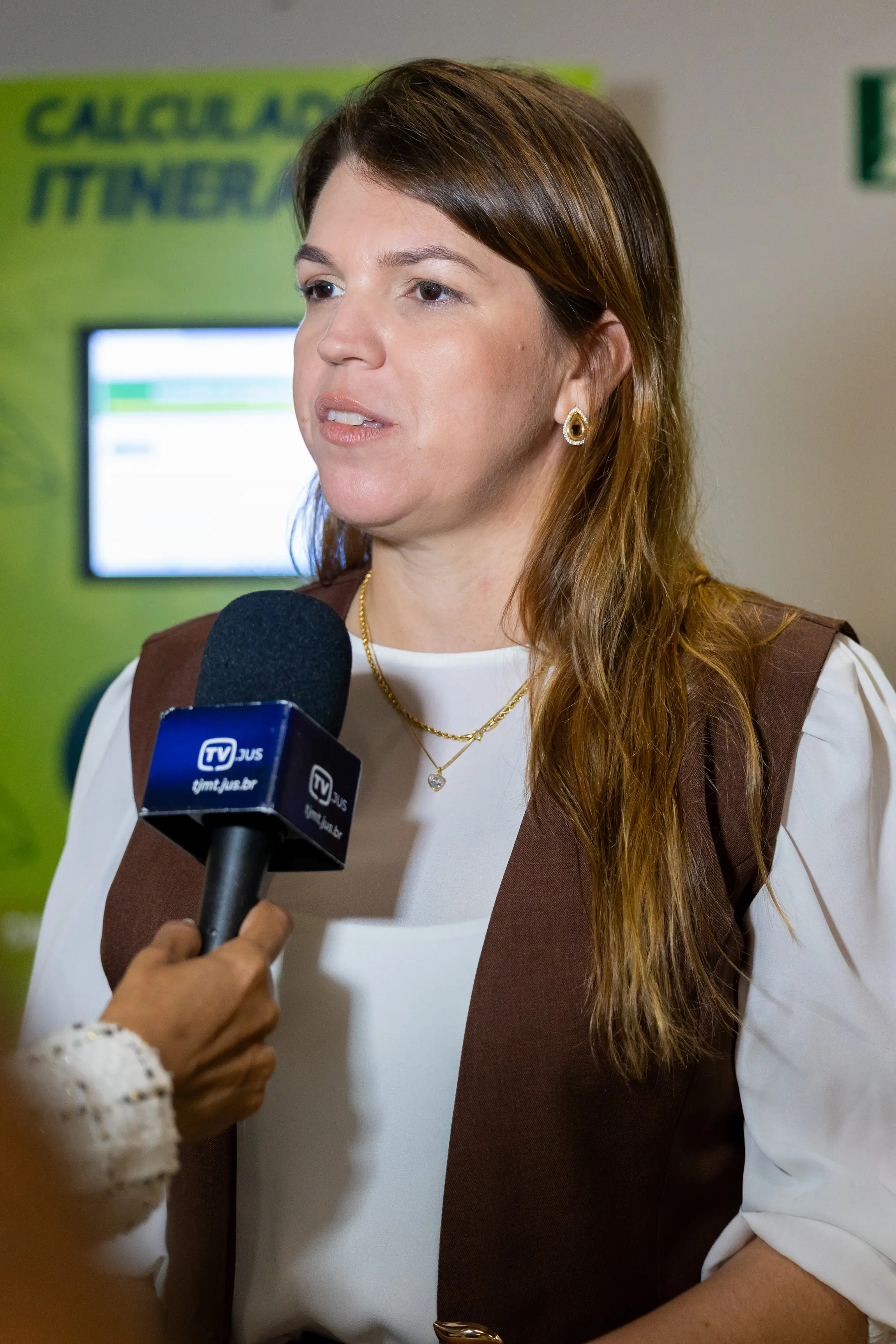 Mulher de pele clara e cabelos castanhos longos, vestindo blusa branca e colete marrom. Ela fala para um microfone com o logo 