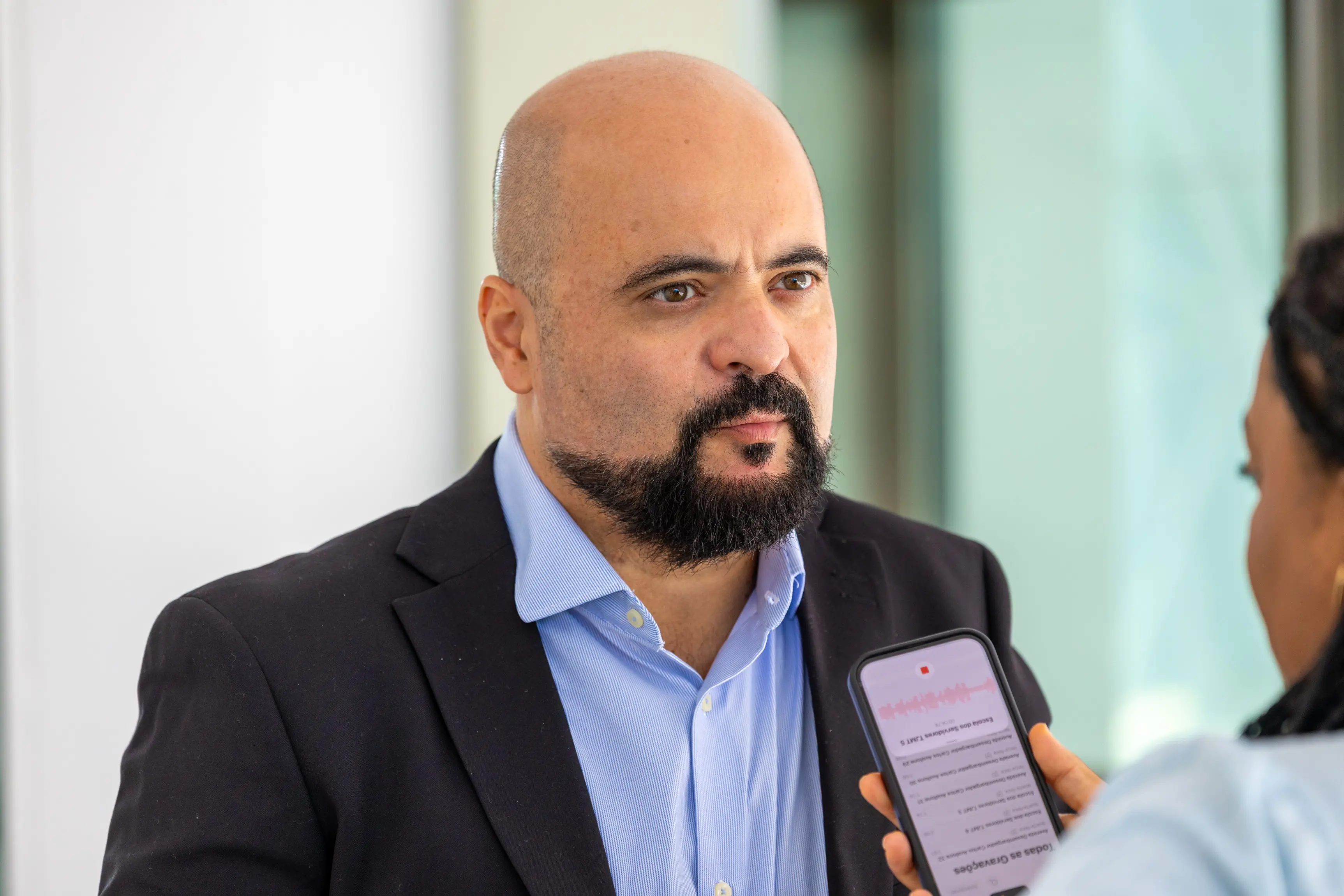 Homem calvo de barba e paletó escuro em close, concedendo entrevista gravada por celular.