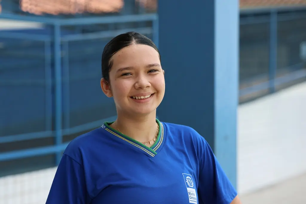 Foto horizontal em plano médio que mostra a estudante Sophia Velazquez sorrindo para a foto, no pátio da escola. Ela é uma adolescente de pele clara, olhos e cabelos escuros, usando uniforme escolar azul.