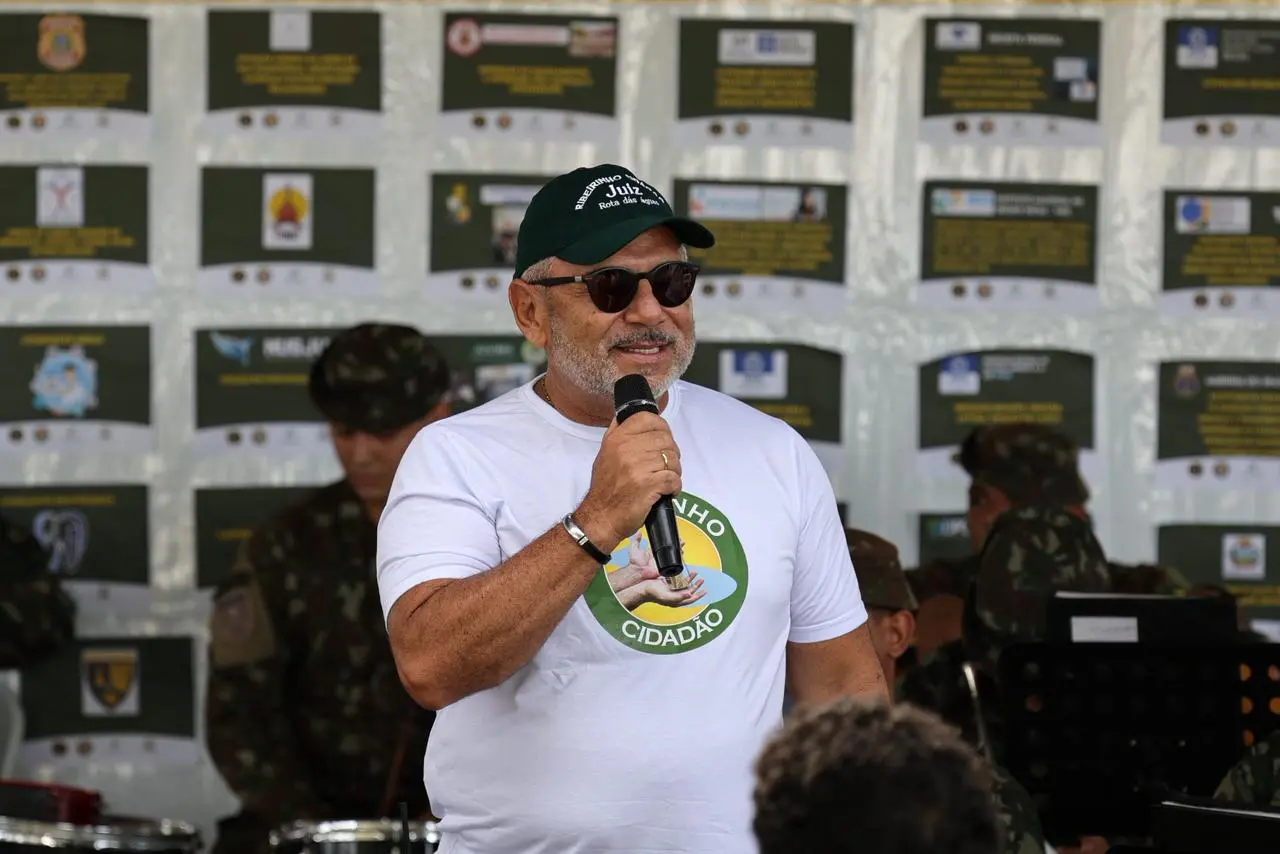 Homem de barba grisalha fala ao microfone. Ele usa camiseta branca com o selo 