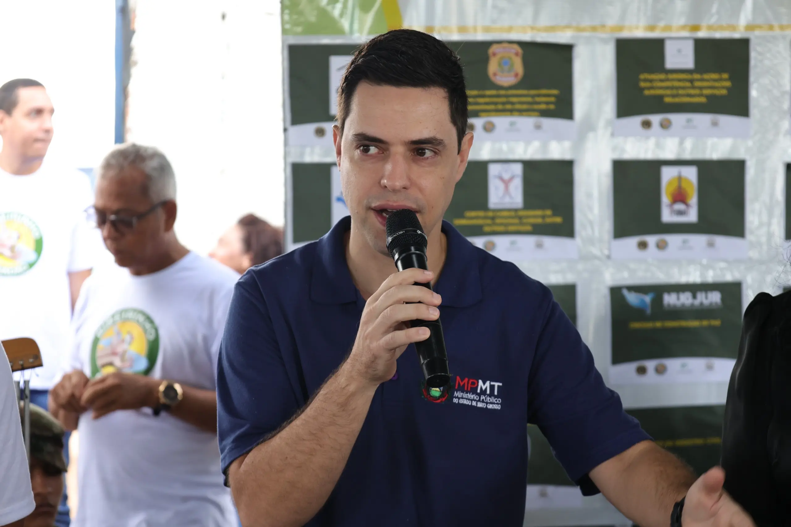 Homem jovem de cabelo curto e pele clara fala ao microfone. Ele veste camisa polo azul marinho com o logotipo do Ministério Público de Mato Grosso (MPMT) no peito.