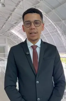 Retrato frontal de um homem jovem, de pele parda e cabelos pretos curtos, usando óculos de grau redondos com armação escura. Ele veste um terno preto clássico, camisa social branca e uma gravata de tom vinho, com um microfone de lapela fixado ao paletó