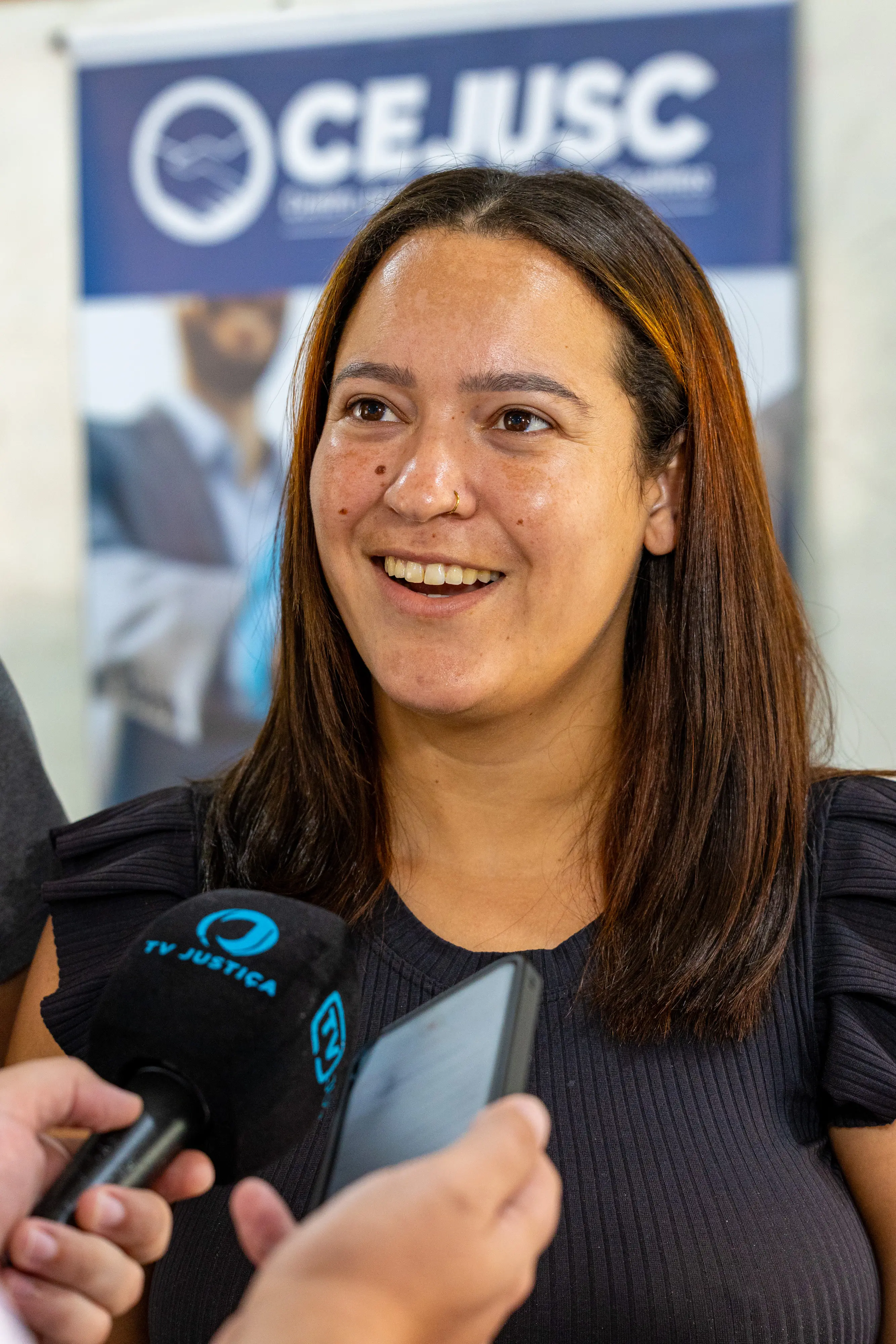 Mulher branca e sorridente, de cabelos castanhos longos e blusa preta, fala ao microfone da TV Justiça e a um celular. Ao fundo, um banner azul com o logotipo do CEJUSC.