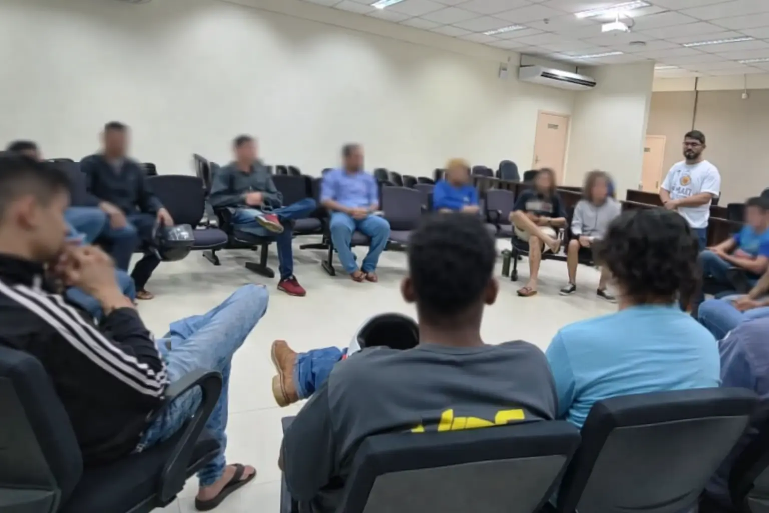 Homens estão sentados em círculo em sala com cadeiras organizadas. Um facilitador em pé conduz a atividade. Os rostos estão desfocados, reforçando privacidade e ambiente de diálogo.