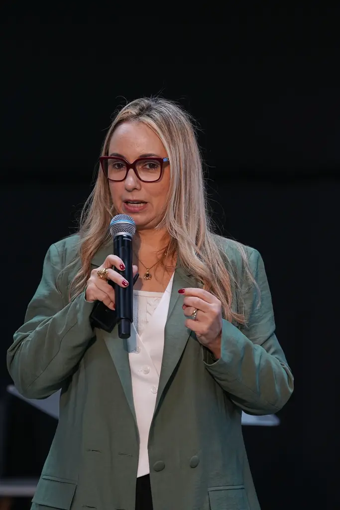 Foto vertical que mostra a juíza Ana Graziela Vaz de Campos em pé, da cintura pra cima, falando ao microfone. ela é uma mulher branca, loira, usando óculos de grau, blusa branca e blazer verde. 