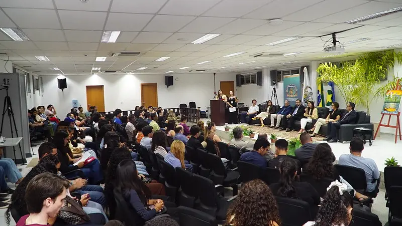 Um auditório lotado recebe o evento Diálogos Acadêmicos, com o público sentado em cadeiras escuras voltadas para um palco lateral onde autoridades debatem em sofás. No palco, destacam-se as bandeiras oficiais, um banner institucional da Esmagis-MT.