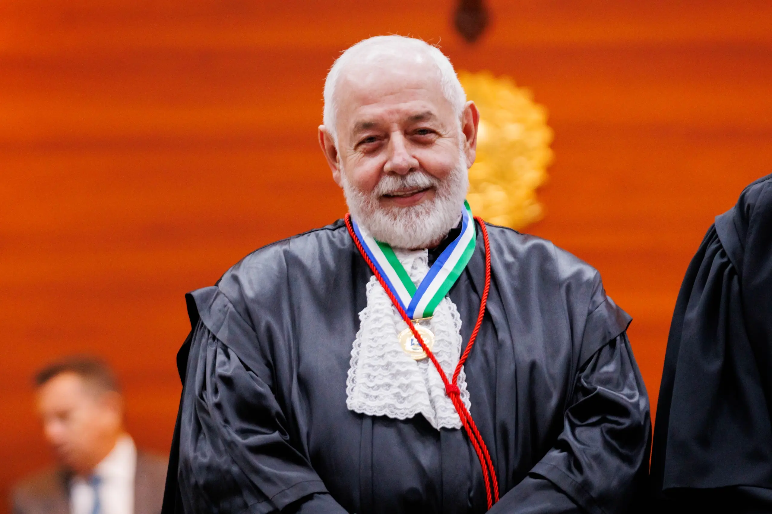 Foto horizontal colorida do presidente José Zuquim sorrindo para foto. Ele é um homem branco, de barba e cabelos brancos. Usa toga de gala preta com babados brancos na frente e medalha envolta do pescoço