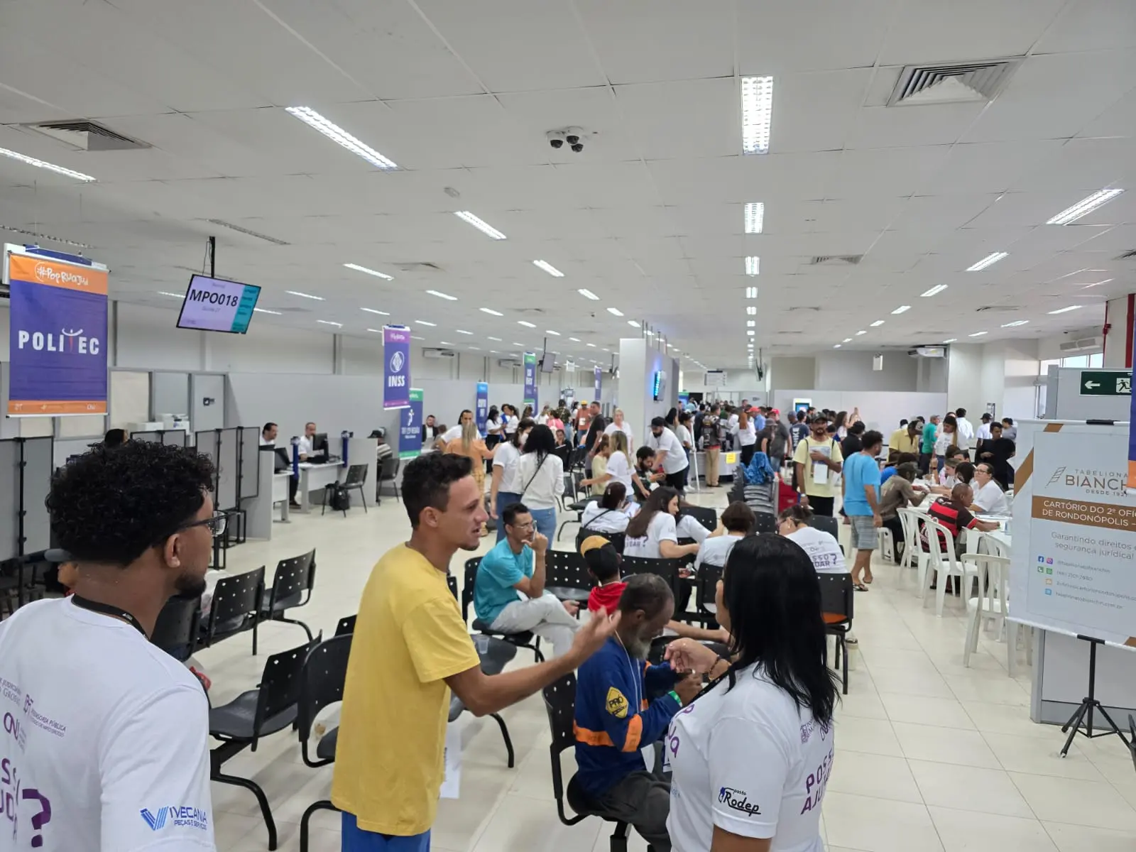 Grande salão com muitas pessoas sendo atendidas em mesas organizadas. Placas indicam serviços como Politec. Participantes conversam, aguardam ou recebem orientação em ambiente amplo e movimentado.