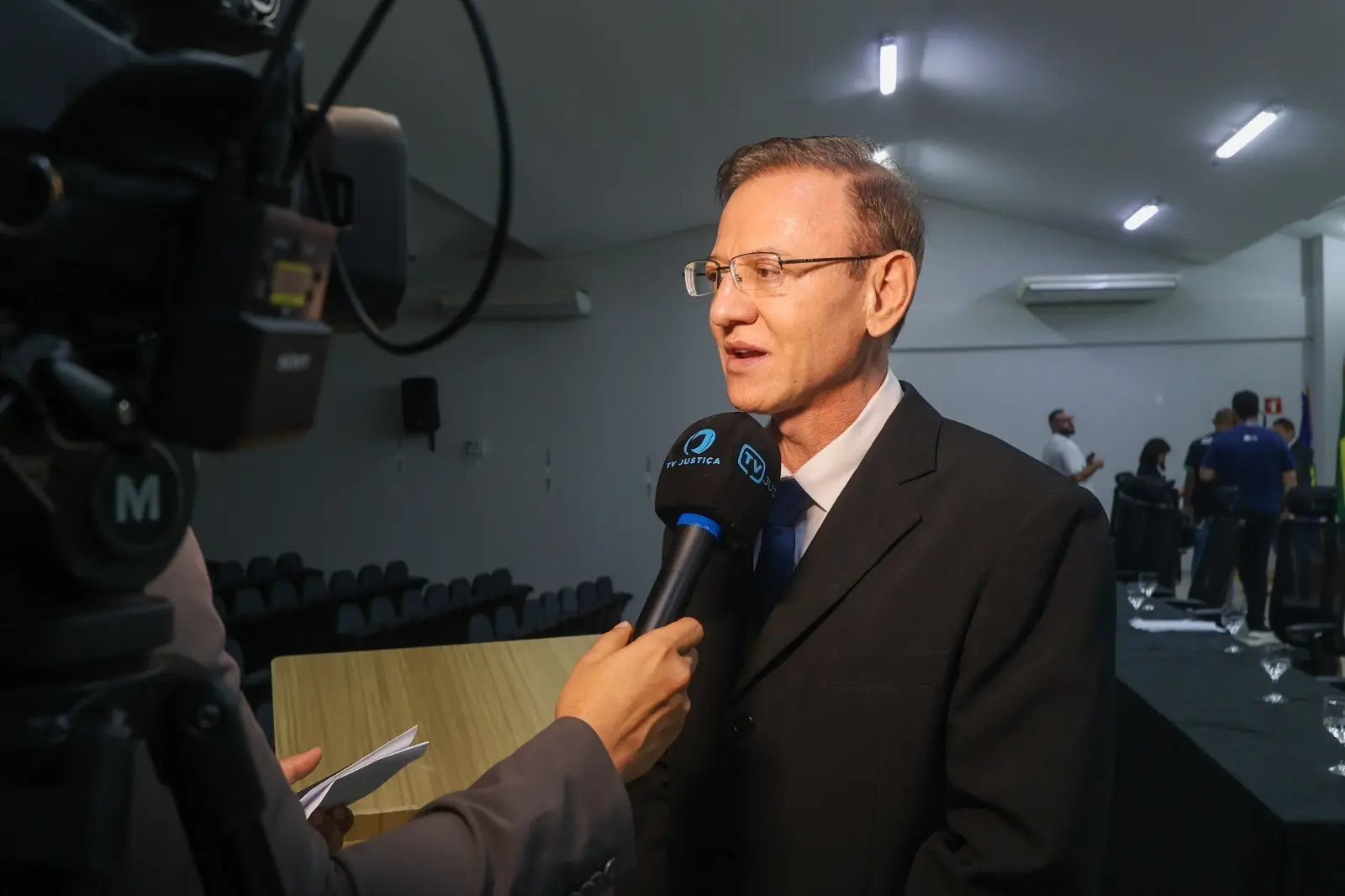 Um homem de terno e óculos concede entrevista. À esquerda, parte de uma câmera profissional e uma mão segurando um microfone com as logomarcas 
