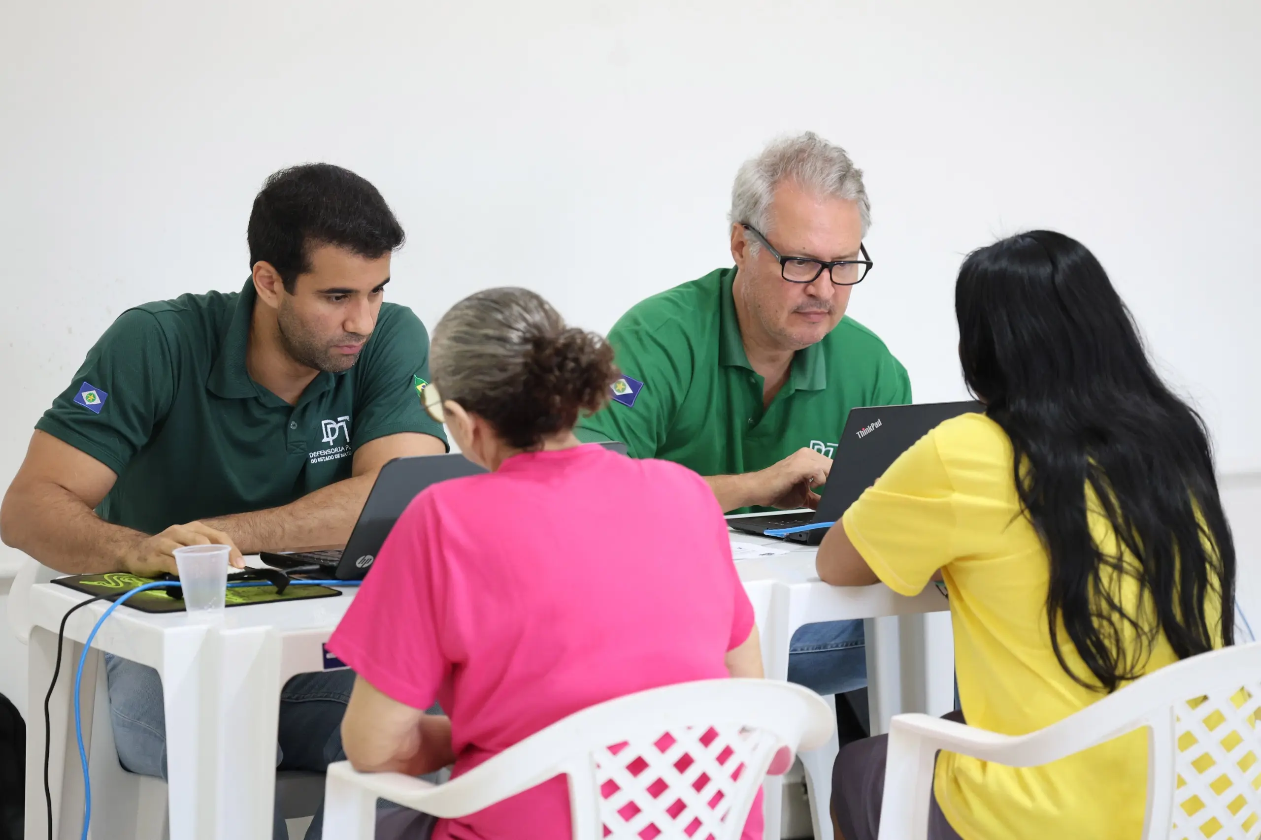 Representantes da Defensoria Pública atendem duas mulheres sentadas à mesa. Os profissionais utilizam notebooks durante o atendimento. As atendidas vestem camisetas nas cores rosa e amarela.