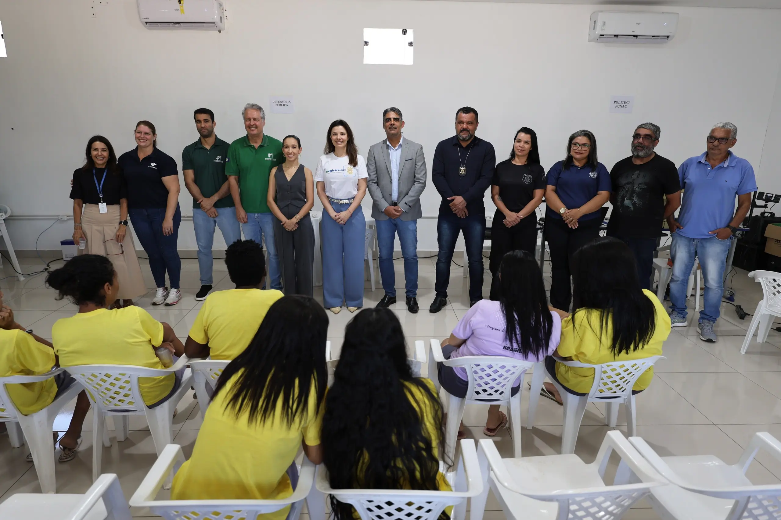 Equipe de instituições parceiras alinhada em frente a pessoas privadas de liberdade sentadas. Os participantes estão em uma sala, com servidores, representantes de órgãos e reeducandas vestindo camisetas amarelas.