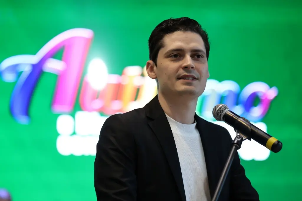 Foto horizontal colorida em plano fechado que mostra o pastor Maik Aquino falando no púlpito. Ele é um homem jovem, branco, de olhos e cabelos castanho escuro, usando camiseta branca e blazer preto. Atrás dele, aparece um telão colorido com a logomarca do evento.
