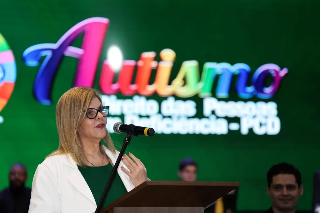 Foto horizontal colorida que mostra a desembargadora Nilza Maria Pôssas de Carvalho do busto pra cima, falando na tribuna. Ela é uma senhora de pele clara, loira, usando vestido verde, blazer branco e óculos de grau de armação preta. Atrás dela há um enorme telão com a logomarca do evento.