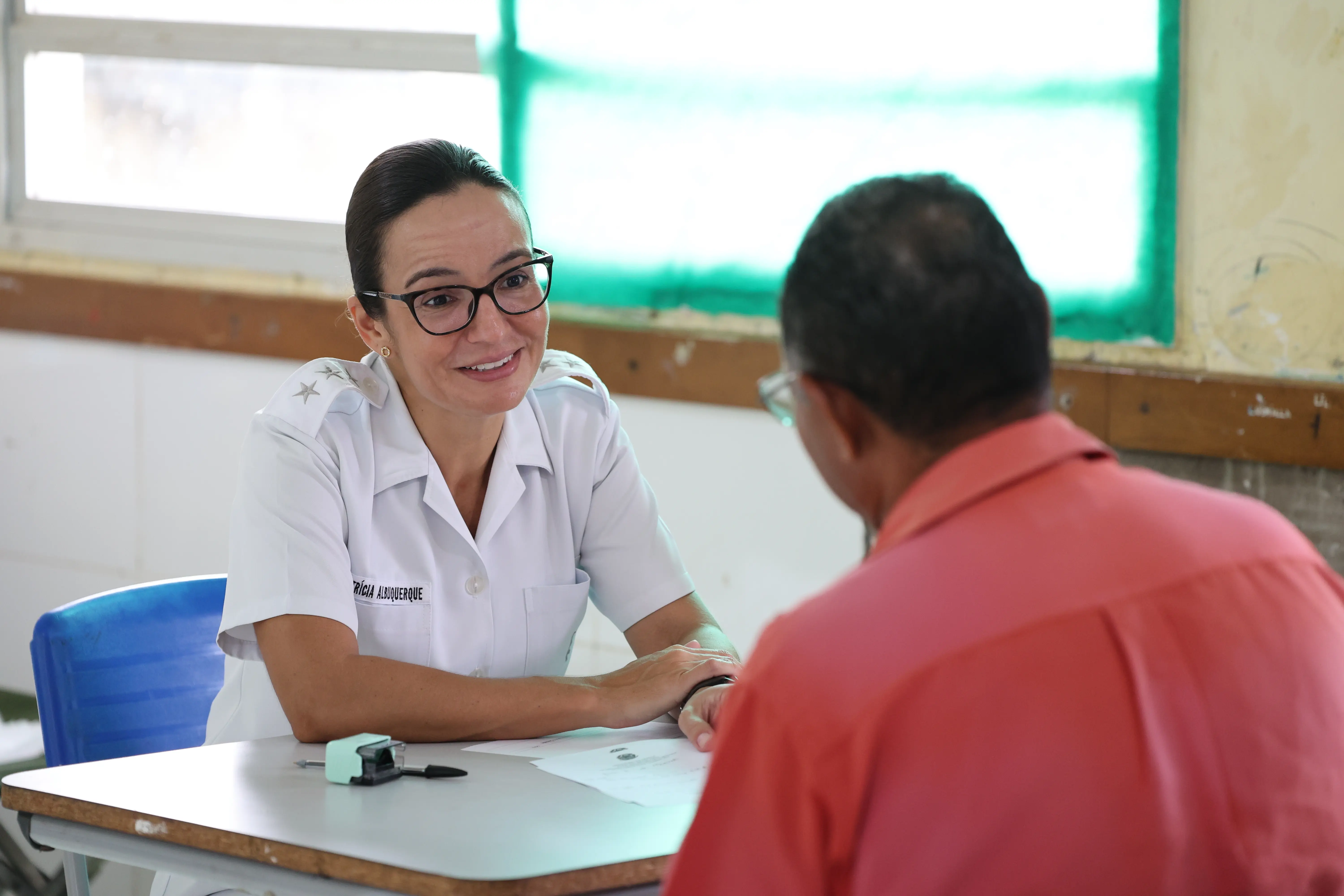 Mulher de óculos e uniforme branco com estrelas nos ombros sorri enquanto atende um homem sentado à sua frente. Eles estão em uma mesa com papéis e um carimbo.