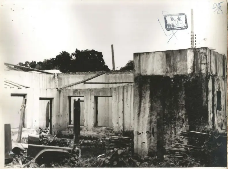 Foto antiga em preto e branco mostra as ruínas da mesma casa. As paredes estão desgastadas e o telhado ruiu, restando apenas algumas vigas de madeira e aberturas de portas e janelas.