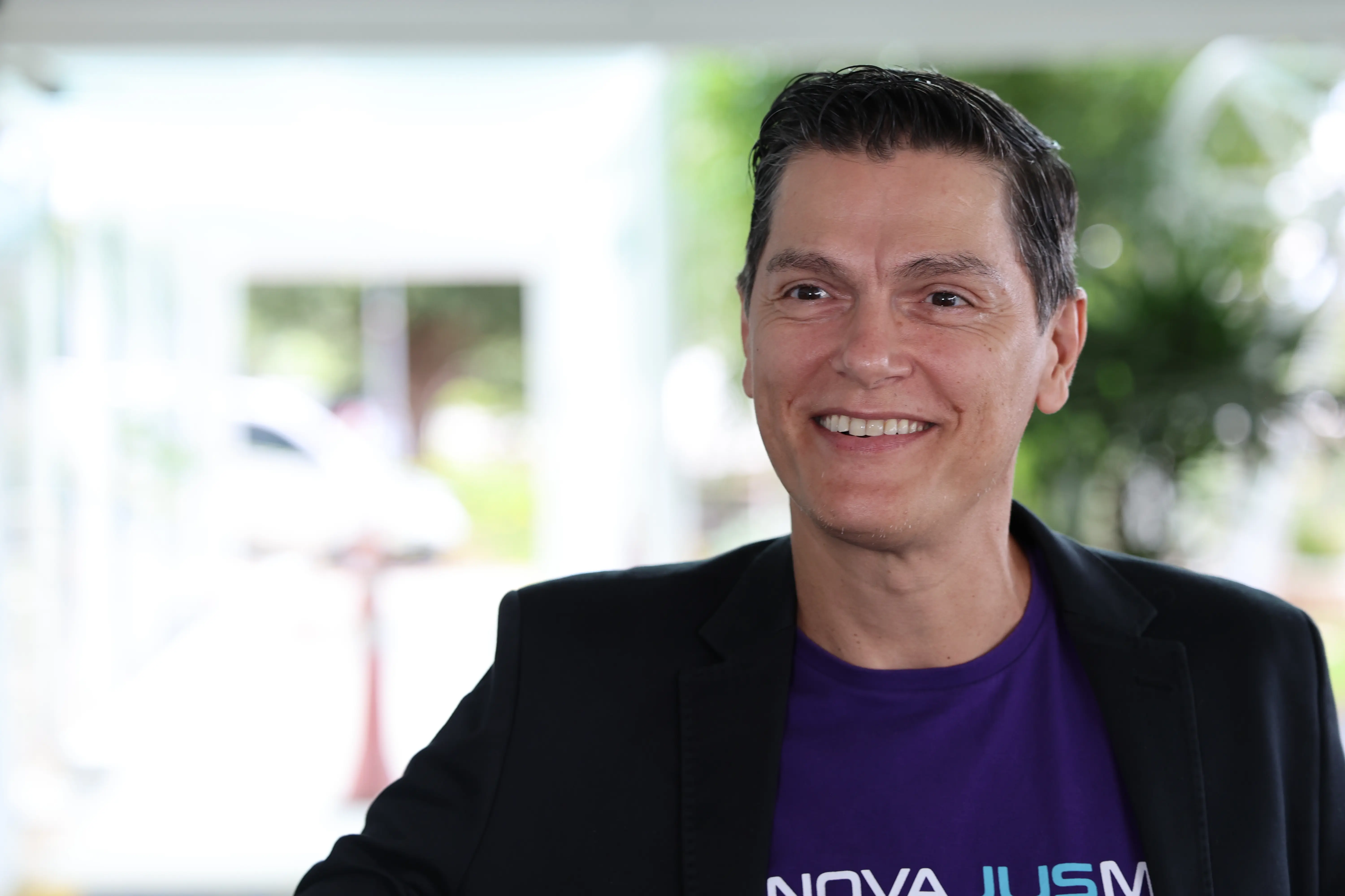 Foto do servidor Thomas Augusto Caetano. Ele está sorrindo e olhando levemente para o lado. Tem cabelos curtos e escuros, veste uma camiseta roxa sob um paletó preto. 