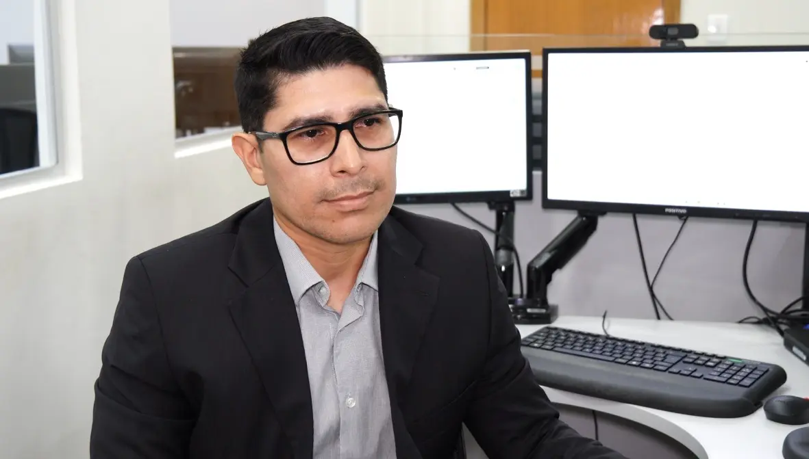 Foto horizontal colorida, em plano fechado, que mostra o gestor de Contabilidade do TJMT, Alexander Hurtado, sentado em seu posto de trabalho, de costas para o computador. Ele é um homem pardo, de olhos e cabelos escuros, usando camisa cinza, terno preto e óculos de grau.