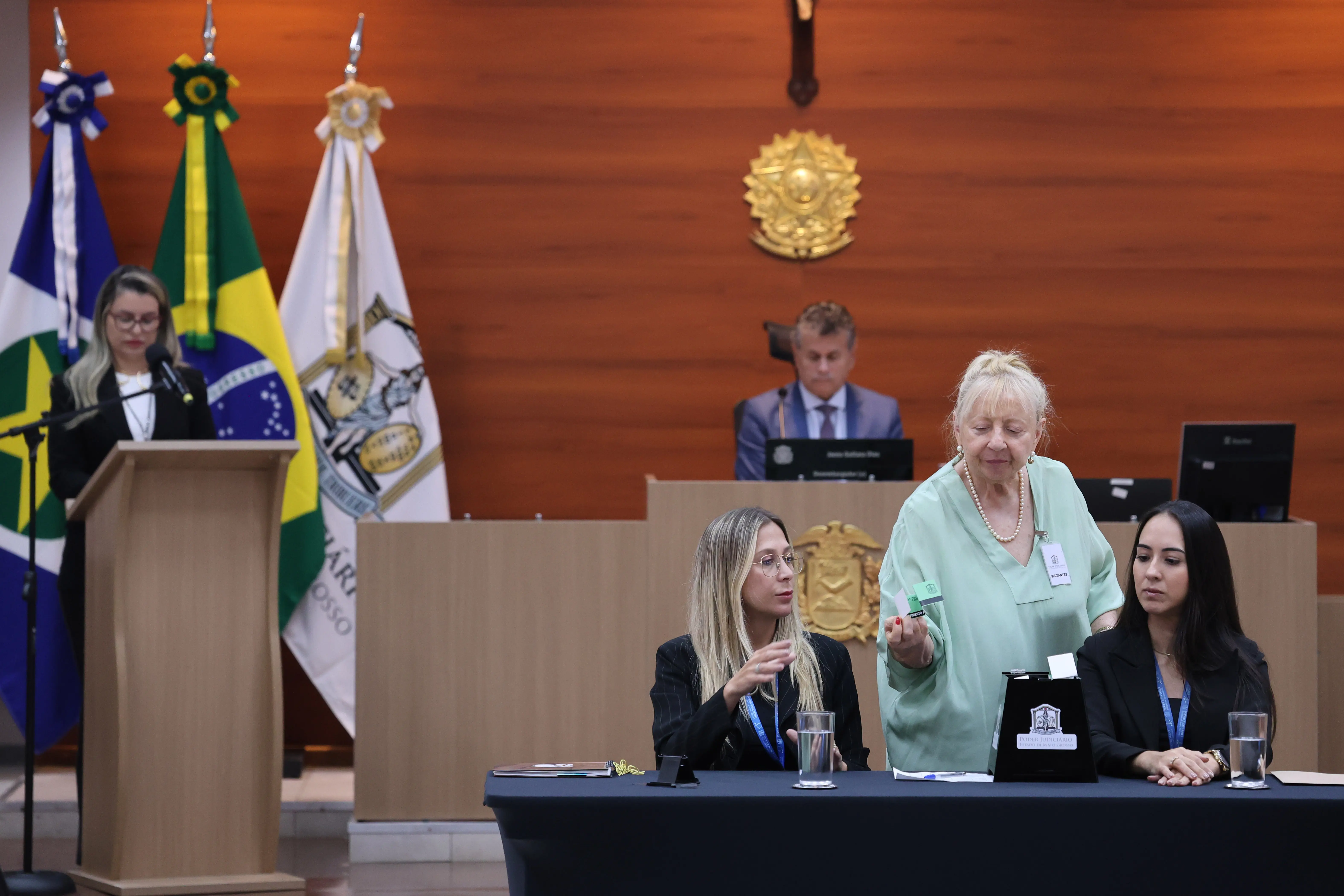Mulher idosa de pele clara, cabelo loiro preso, blusa verde-clara e colar de pérolas, entrega papel. Ao lado, duas jovens de pele clara, blazers pretos; uma loira de óculos, outra morena de cabelo liso.