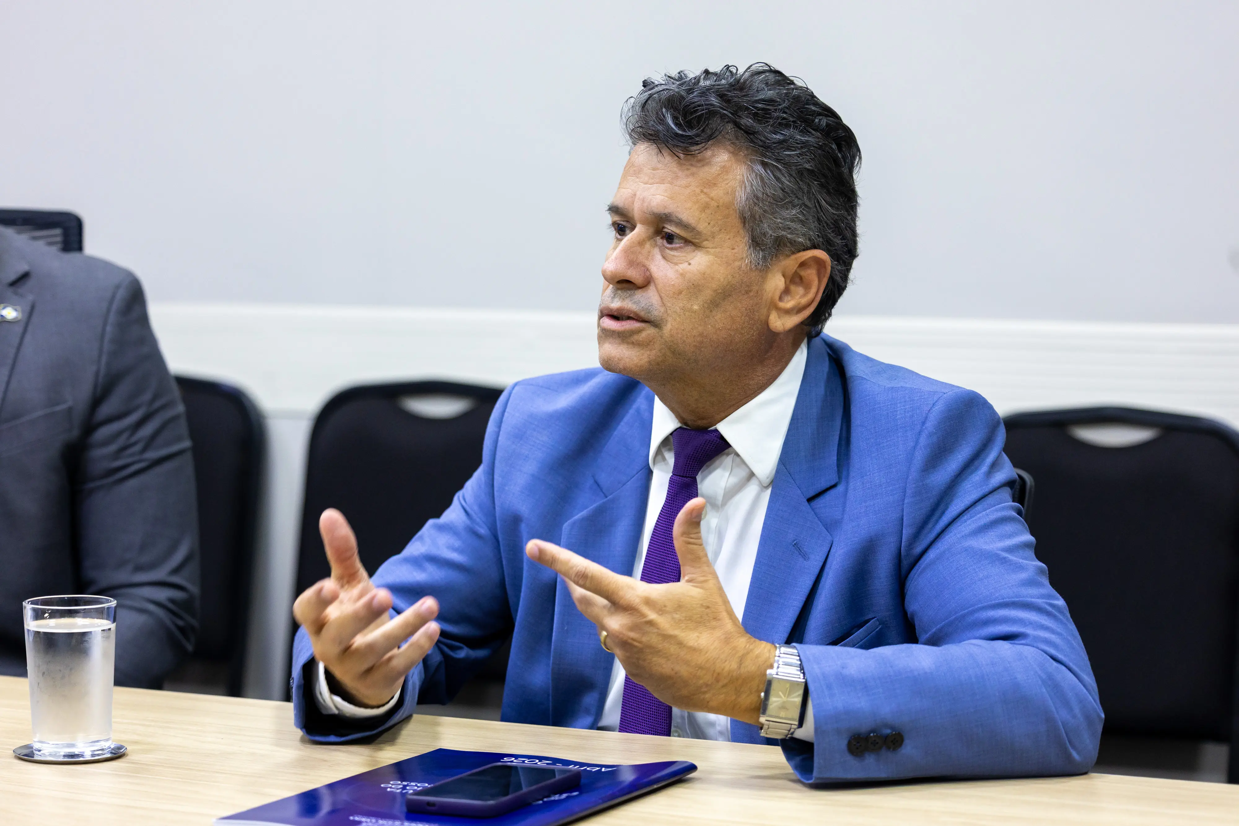 Homem de terno azul e gravata roxa gesticula com as mãos enquanto fala em uma reunião. Ele está sentado à mesa com um copo de água, um celular e uma pasta à sua frente.
