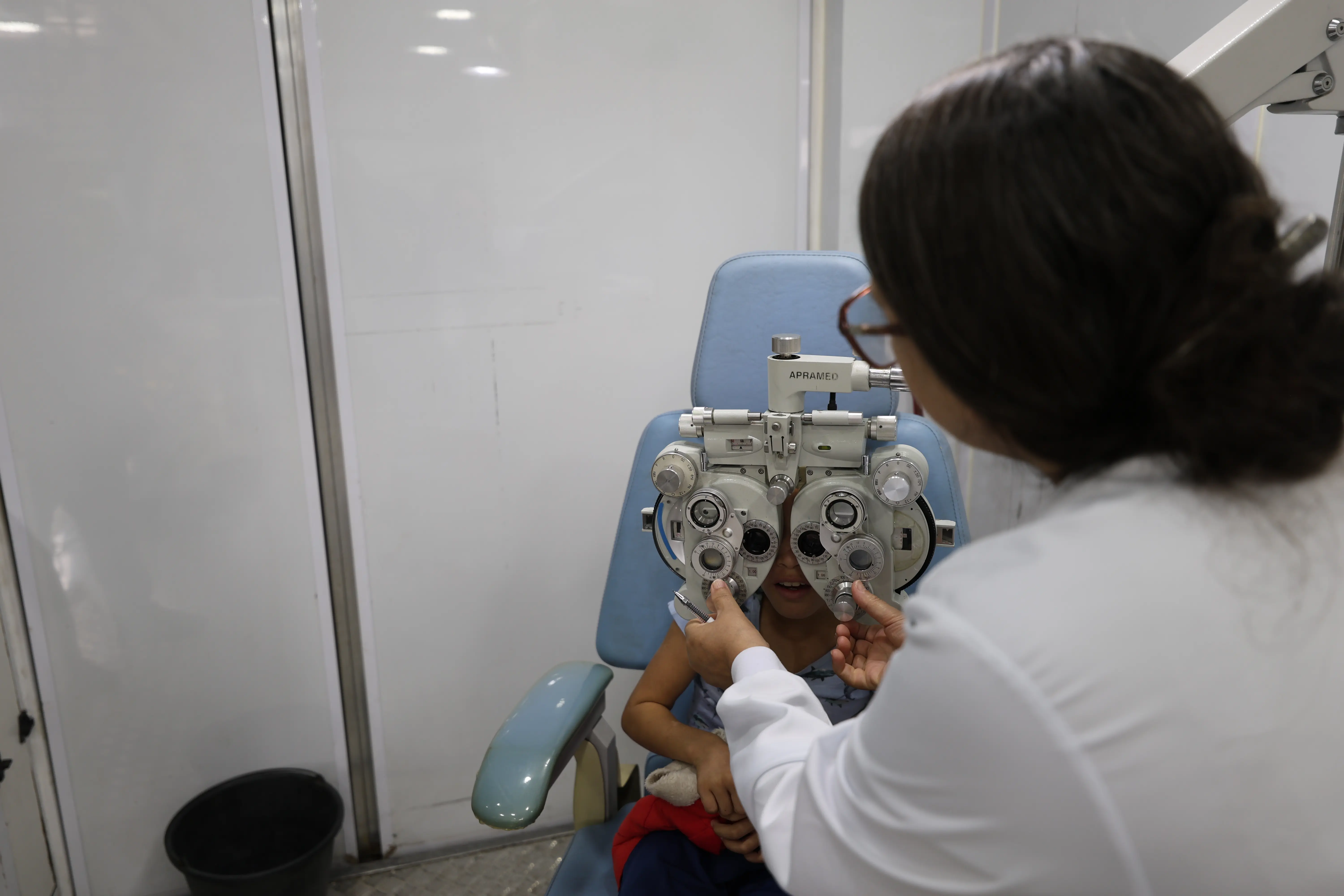Uma criança sentada realiza exame oftalmológico. Uma profissional de jaleco branco ajusta um grande equipamento de refração (refrator) posicionado à frente dos olhos do paciente.