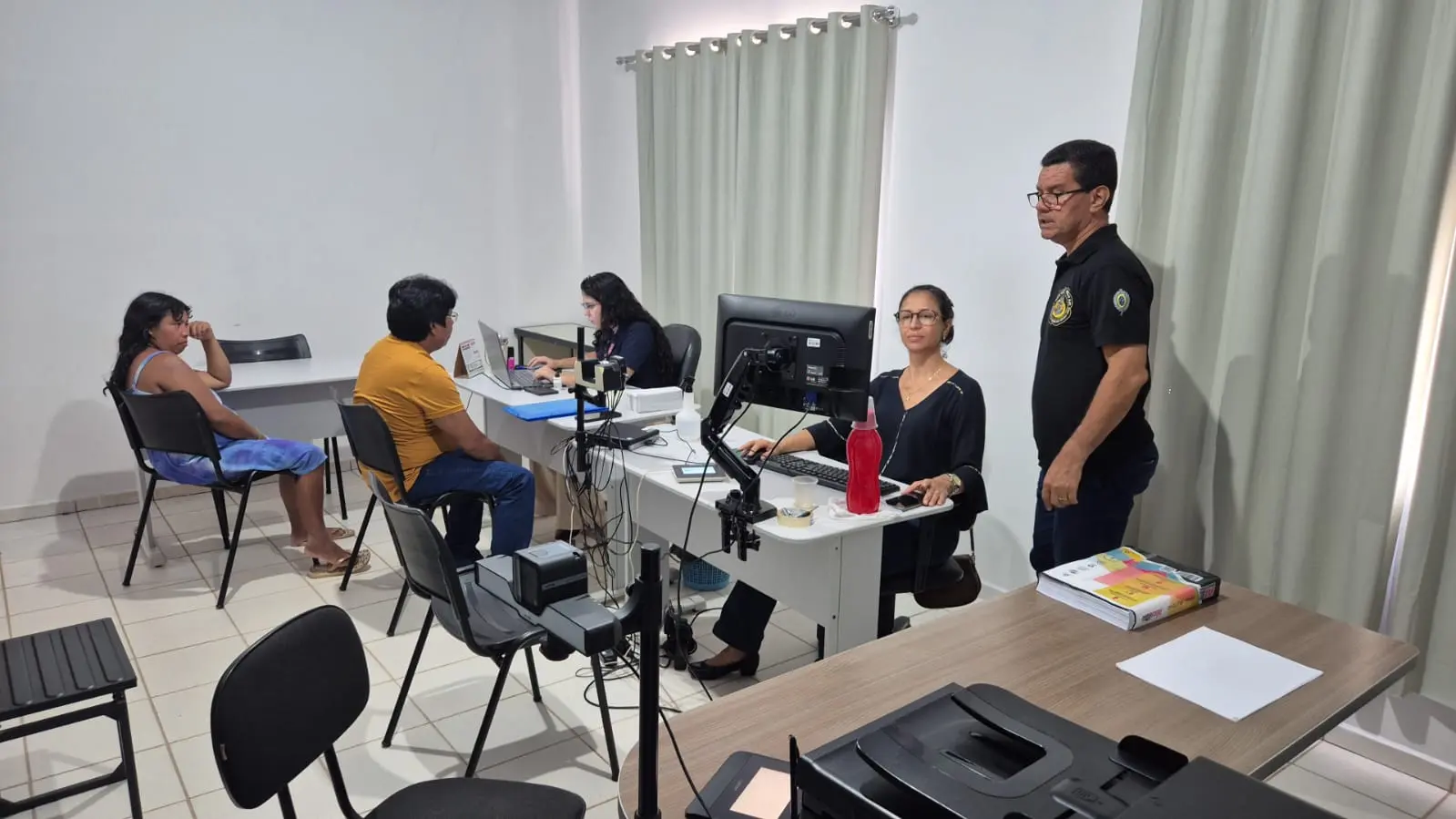 Em uma sala de atendimento, parceiros do Registre-se realizam atendimentos à população indígena. Servidores estão posicionados em mesas com computadores e equipamentos de coleta de dados, enquanto a população aguarda e são atendidos individualmente