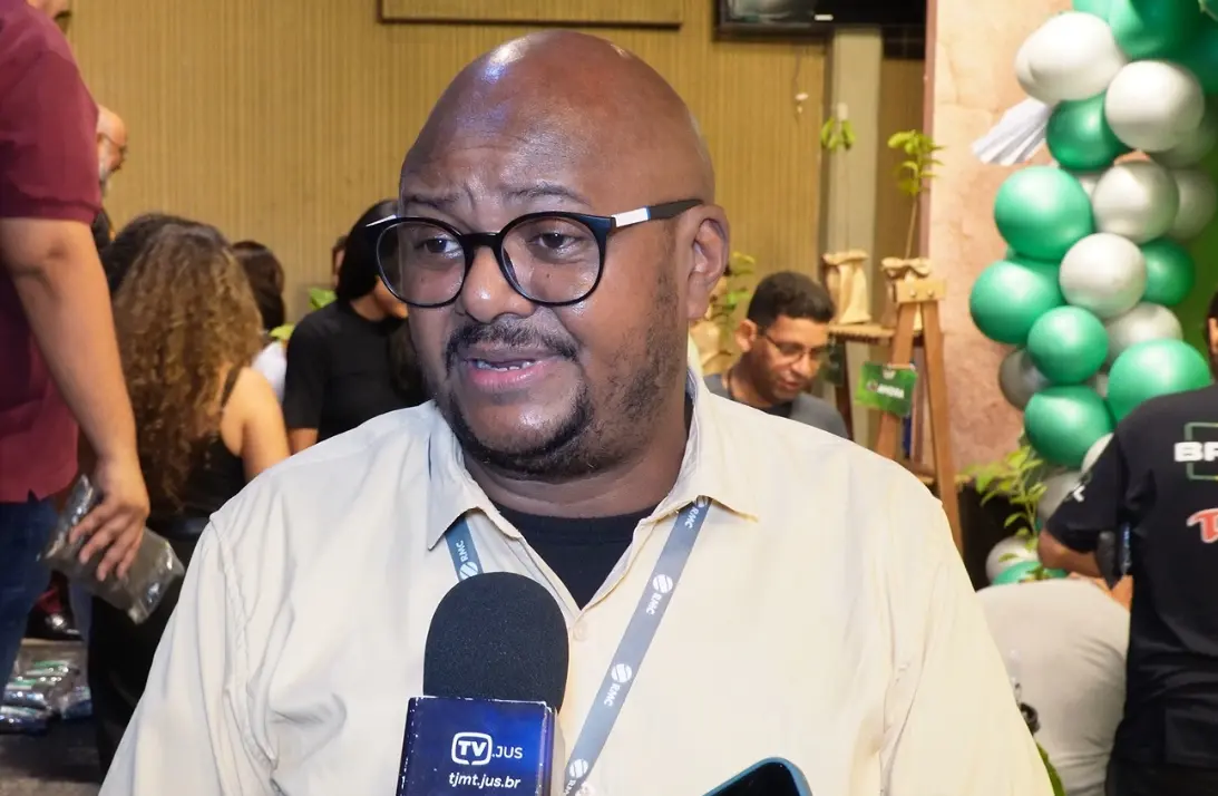 Homem negro de óculos e camisa bege fala ao microfone da TV JUS. Ao fundo, um arco de balões verdes e brancos e pessoas circulando.