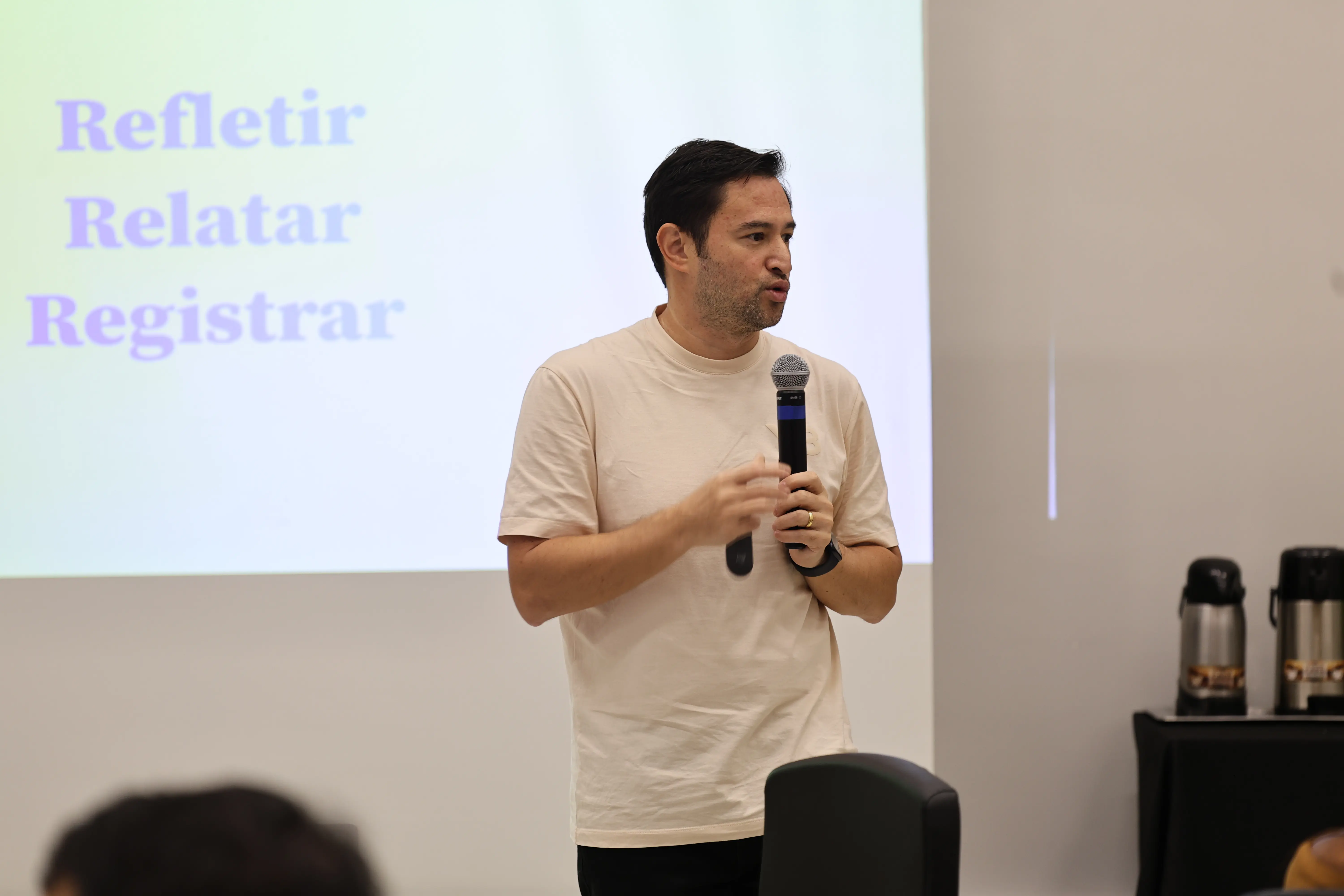 Homem jovem de camiseta clara fala ao microfone diante de uma tela de projeção com as palavras 