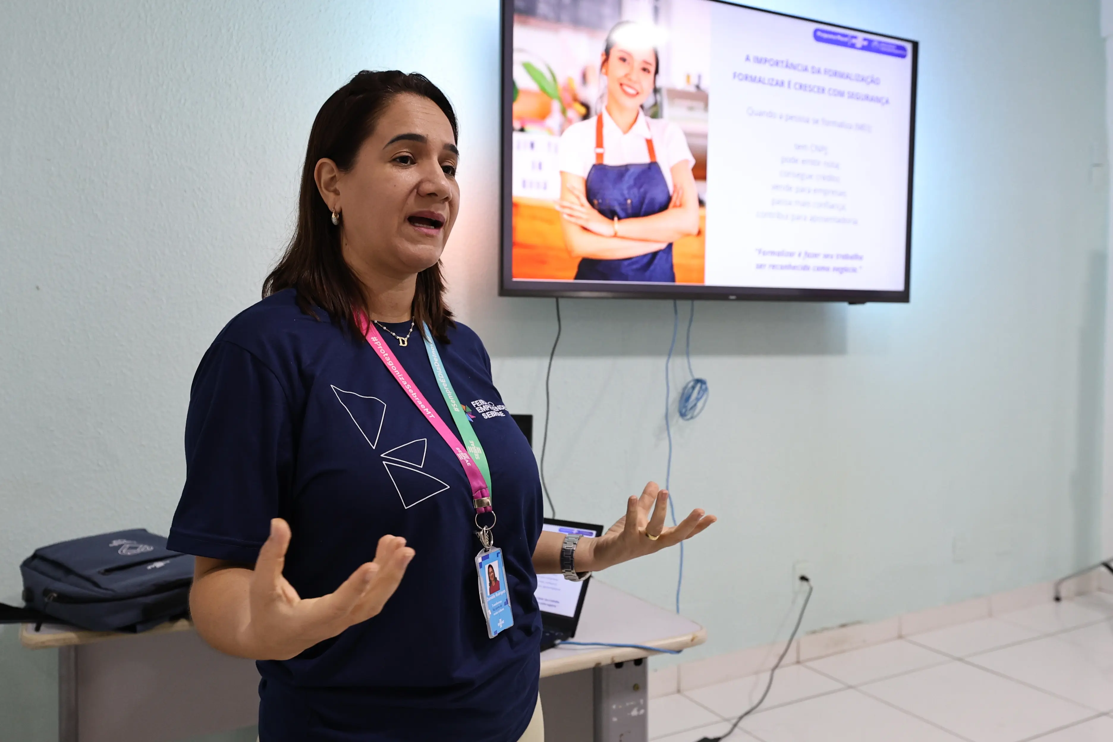 Mulher de camiseta azul marinho e crachá gesticula à frente de uma tela de TV que exibe uma apresentação sobre formalização de negócios. Ambiente de sala de aula.