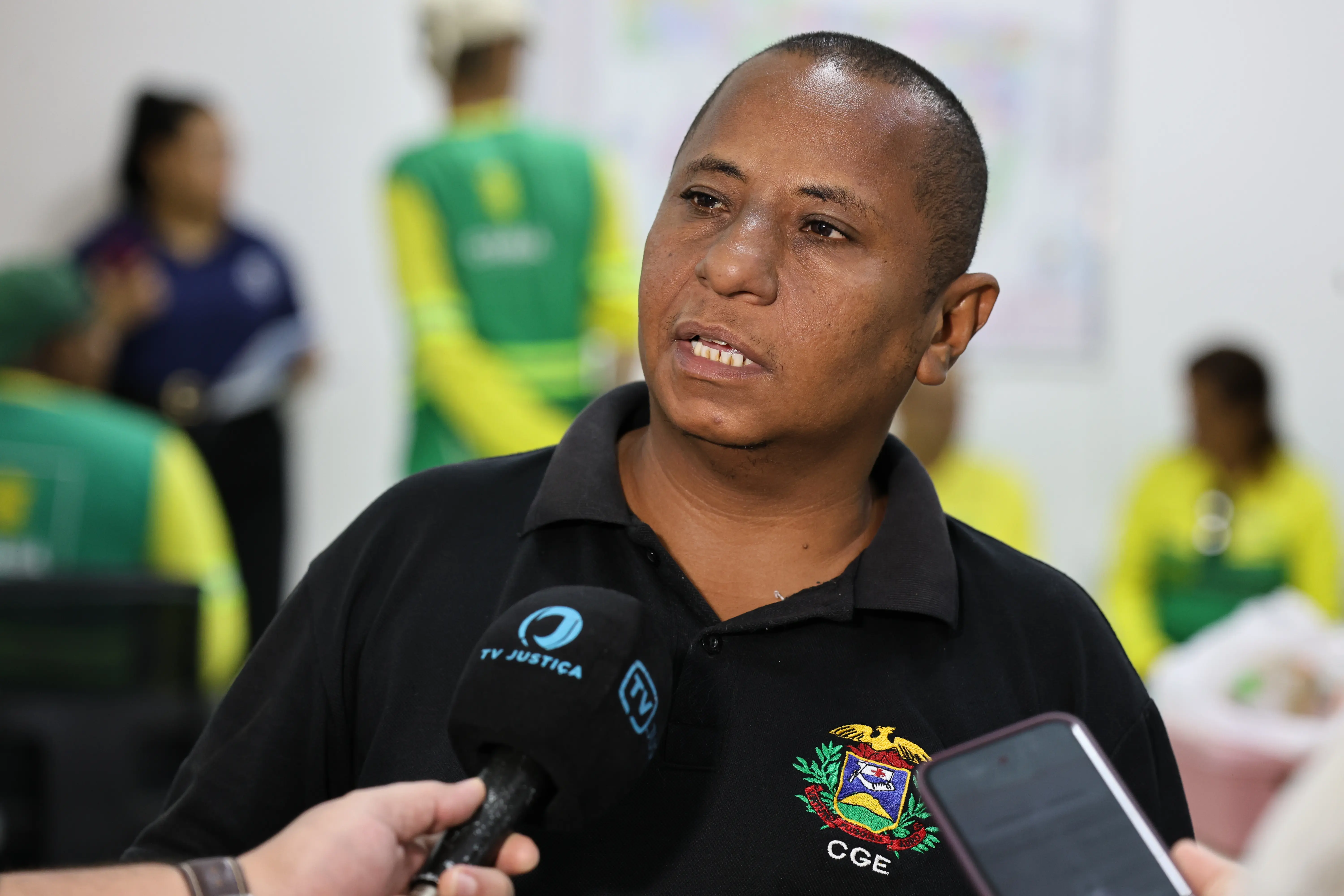 Homem pardo de cabelo curto, vestindo camisa polo preta com brasão e a sigla CGE. Ele fala diante de um microfone da TV Justiça. Ao fundo, pessoas com uniformes verdes e amarelos.