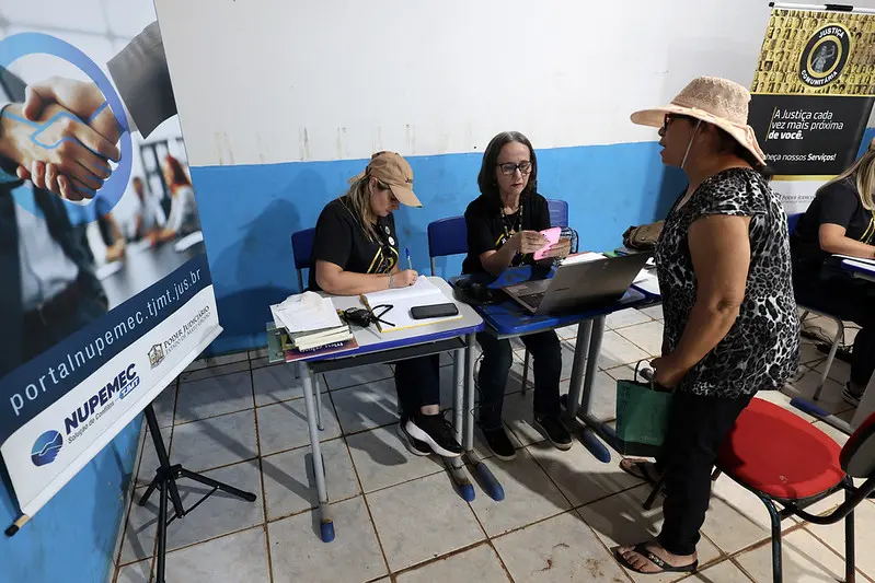 Foto horizontal colorida, em plano aberto, que mostra a senhora Rita Maria Soares em pé, sendo atendida por duas servidoras da Justiça Comunitária, em uma sala de aula, onde ocorreu o mutirão Justiça em Ação. No lado esquerdo da foto, aparece um banner do Cejusc. 