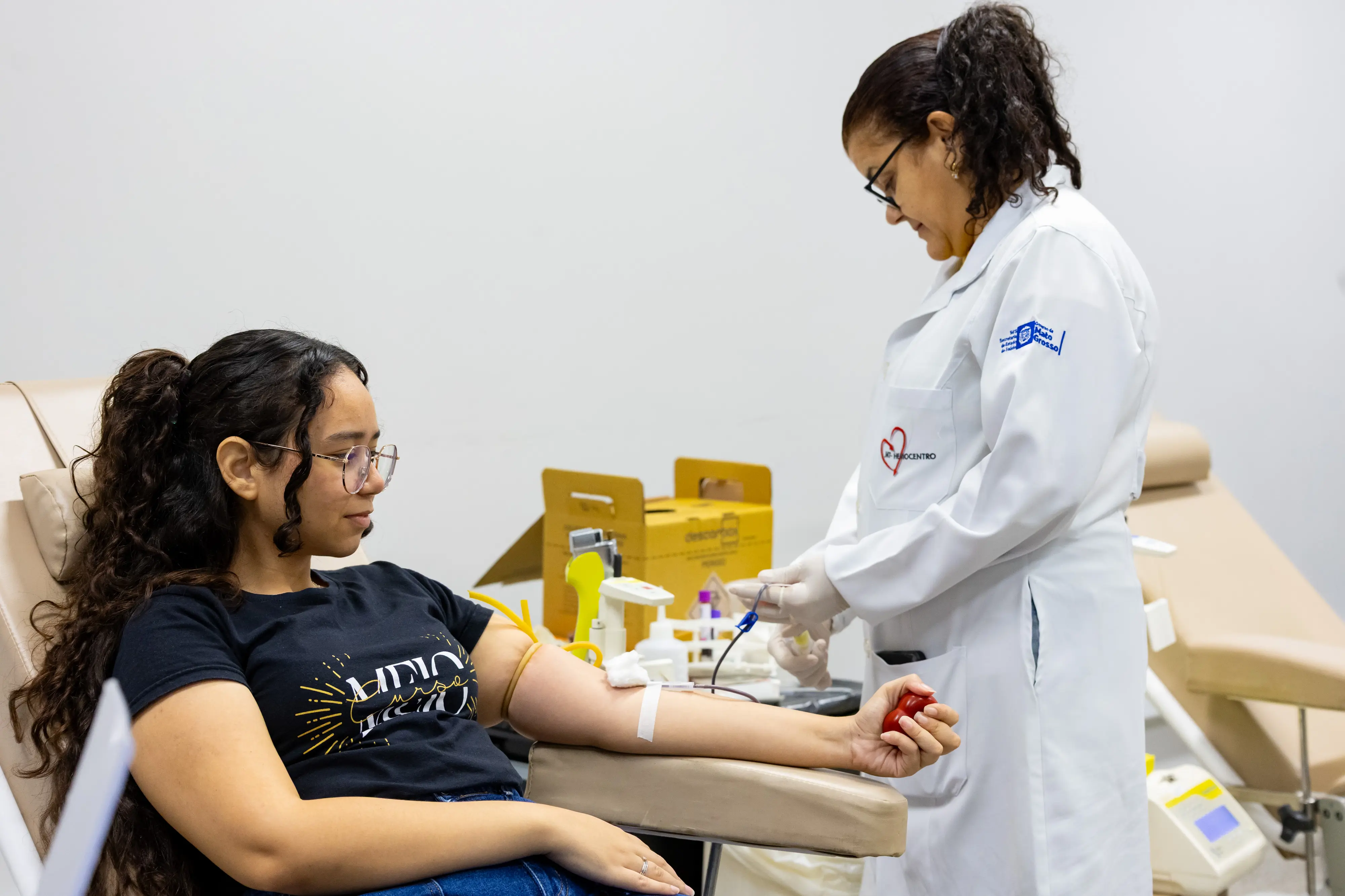 Mulher de óculos, sentada para doar sangue, enquanto uma profissional de saúde do MT-Hemocentro organiza os materiais.