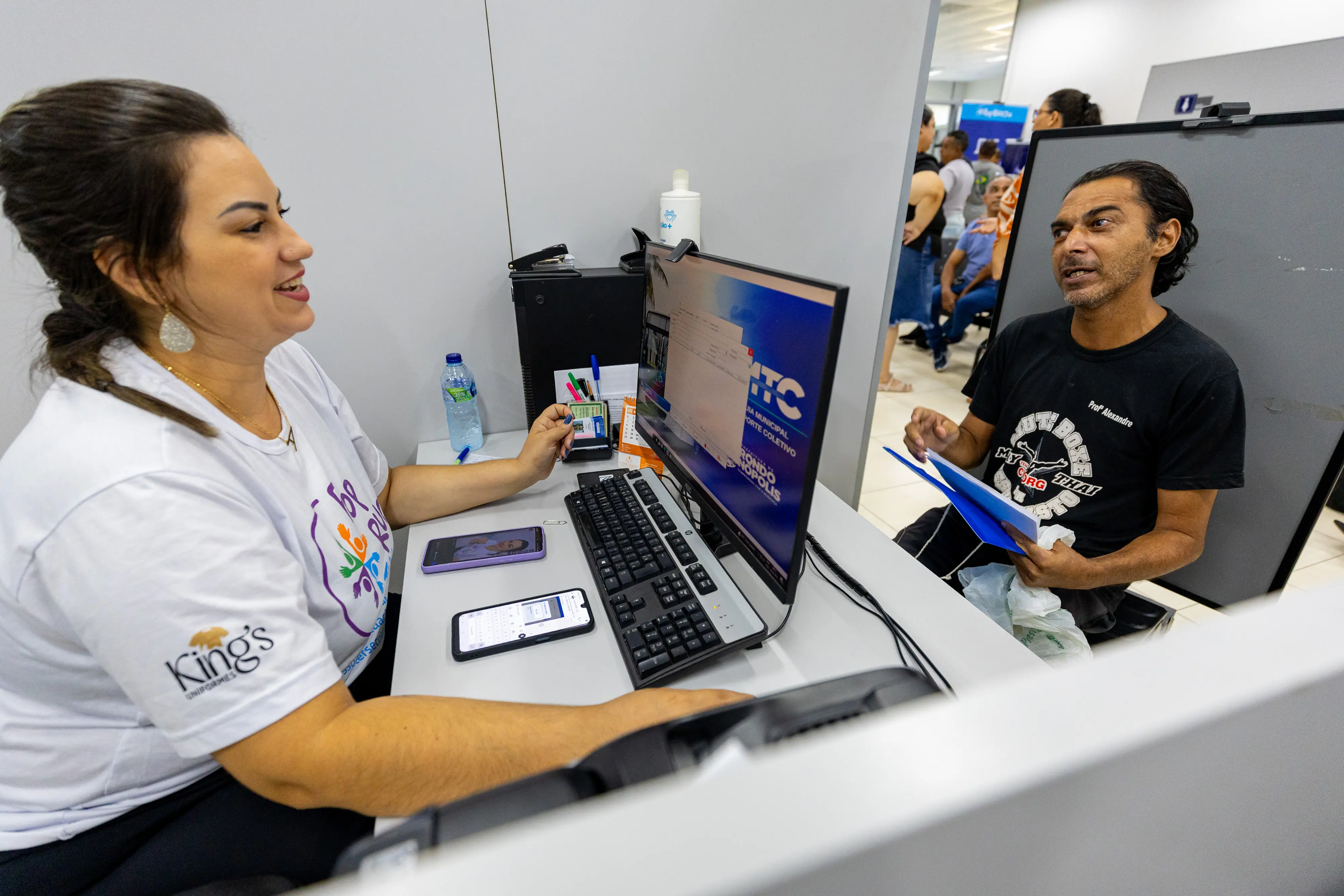 Mulher em guichê de atendimento conversa com homem sentado à sua frente. Entre eles, um computador mostra o sistema. O ambiente é amplo e sugere uma ação de serviços públicos.