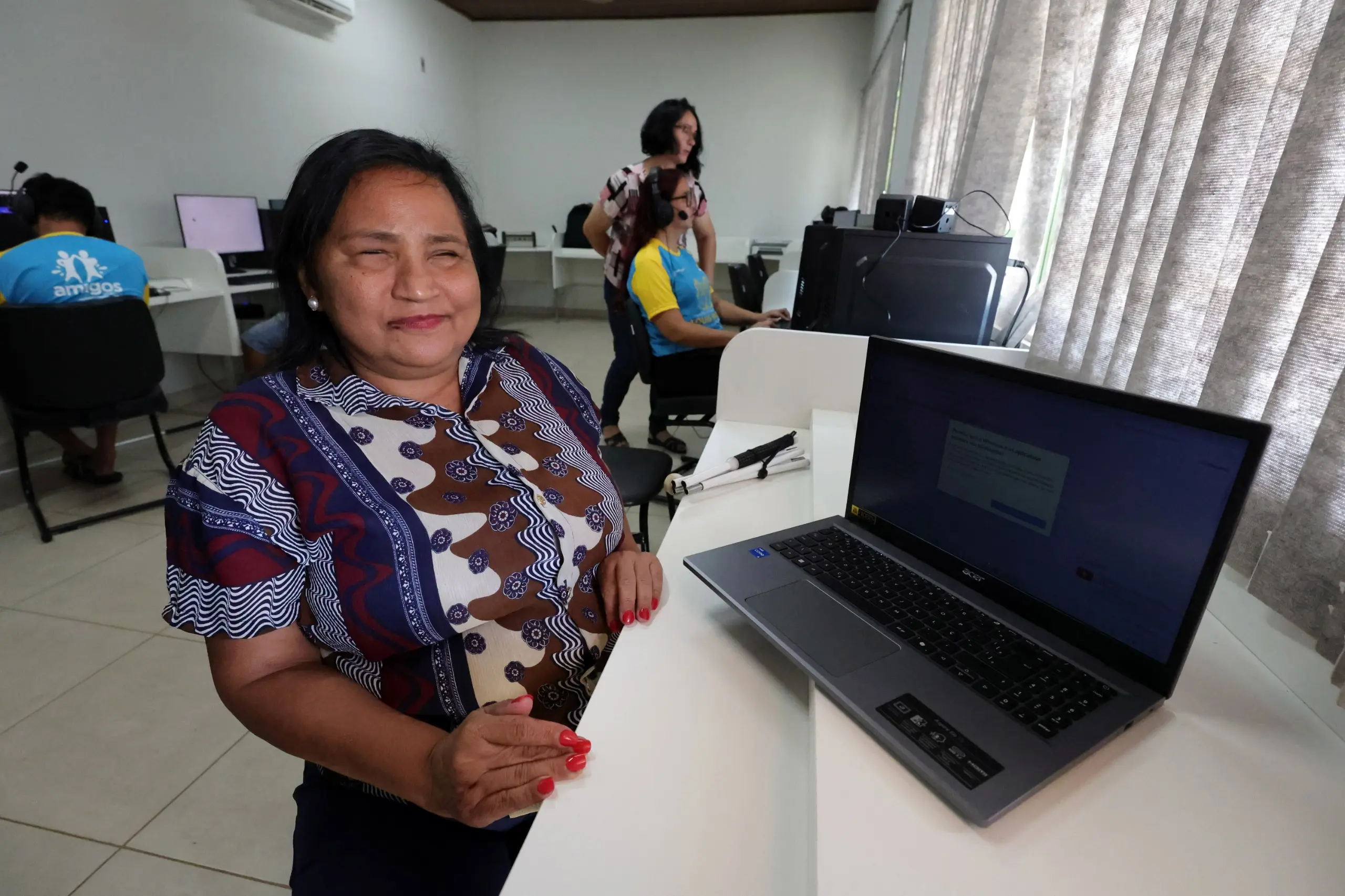 Mulher parda sorridente, com cabelos pretos e blusa estampada, está em pé ao lado de um notebook aberto sobre uma bancada branca. Ao fundo, uma sala com computadores e outras pessoas.