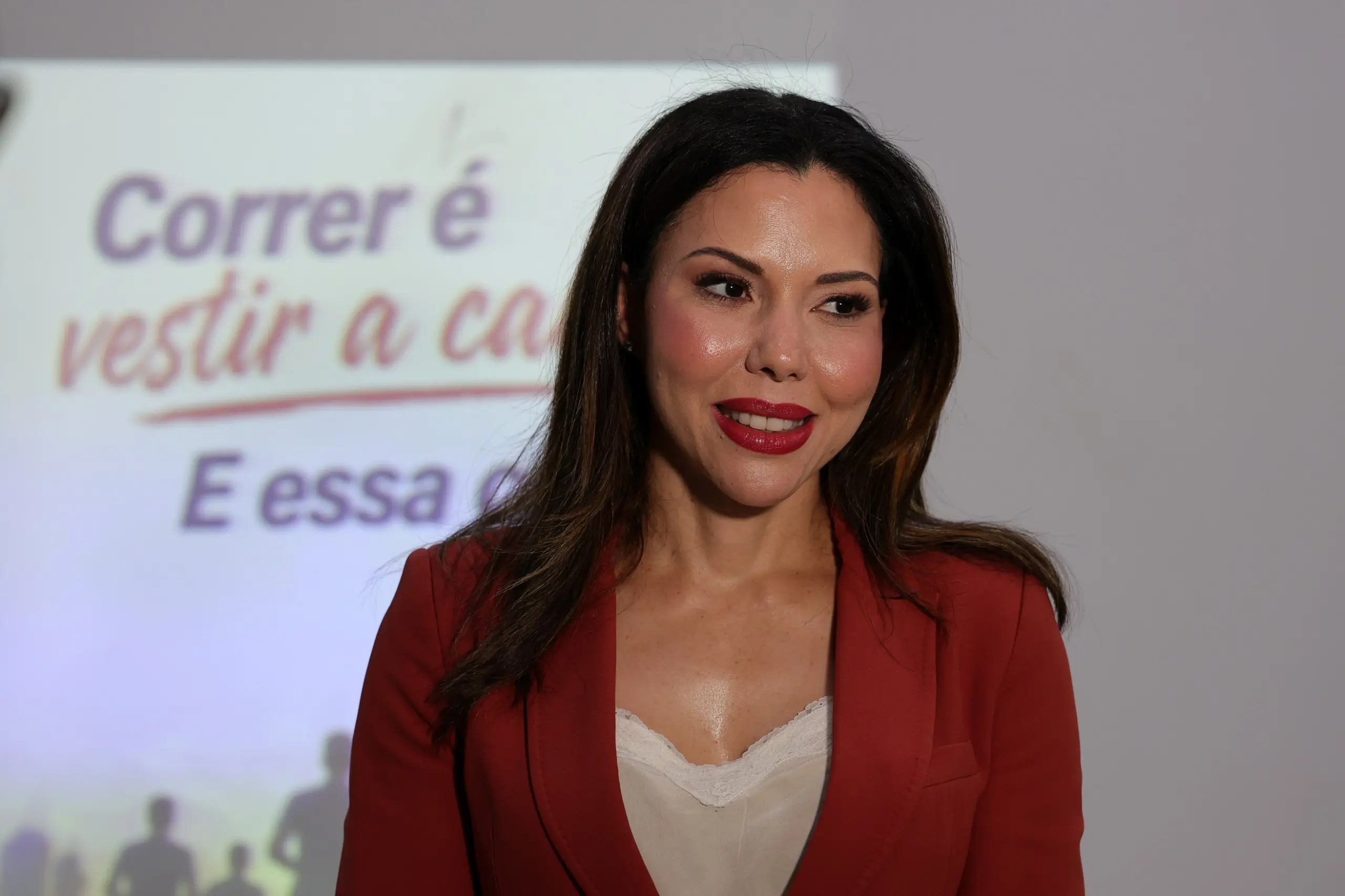 Mulher branca de cabelos longos e castanhos, batom vermelho e blazer vermelho sobre blusa clara. Ao fundo desfocado, uma projeção com texto em português começa com a palavra Correr.