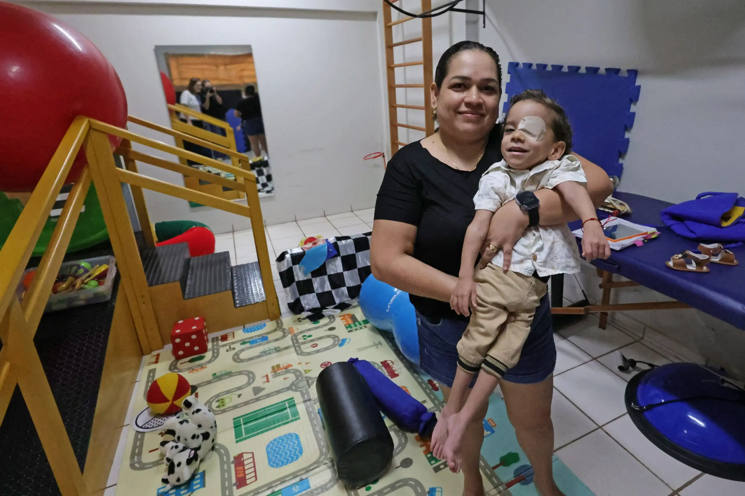 Mulher sorridente segura no colo um menino com tampão no olho esquerdo. Eles estão em uma sala de fisioterapia infantil com tatame colorido, brinquedos, bola de pilates e escada de canto.
