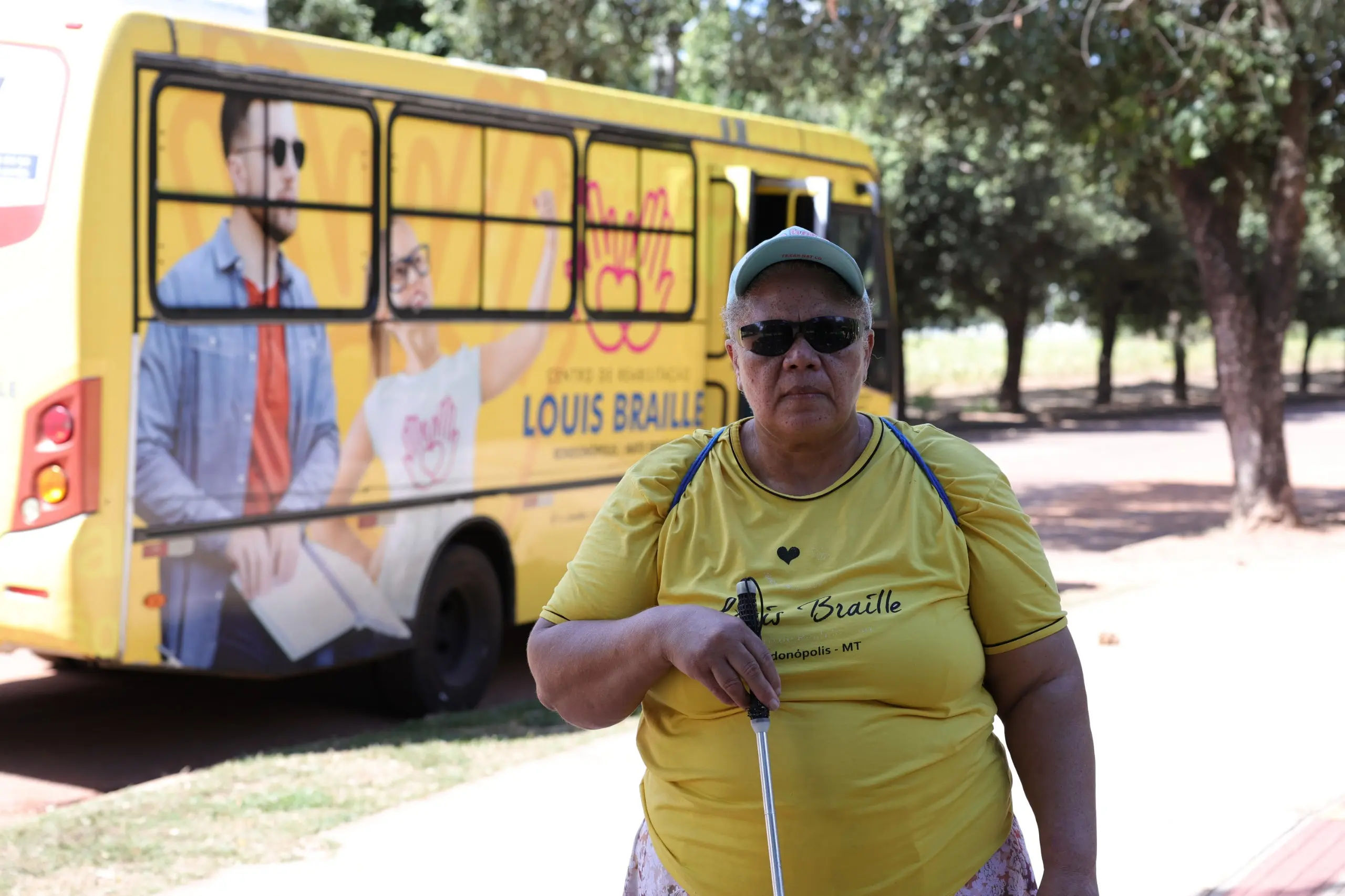 Mulher negra de óculos escuros, boné claro e camiseta amarela segura uma bengala guia. Ao fundo, um ônibus amarelo com imagens de pessoas e a inscrição Centro de Reabilitação Louis Braille.