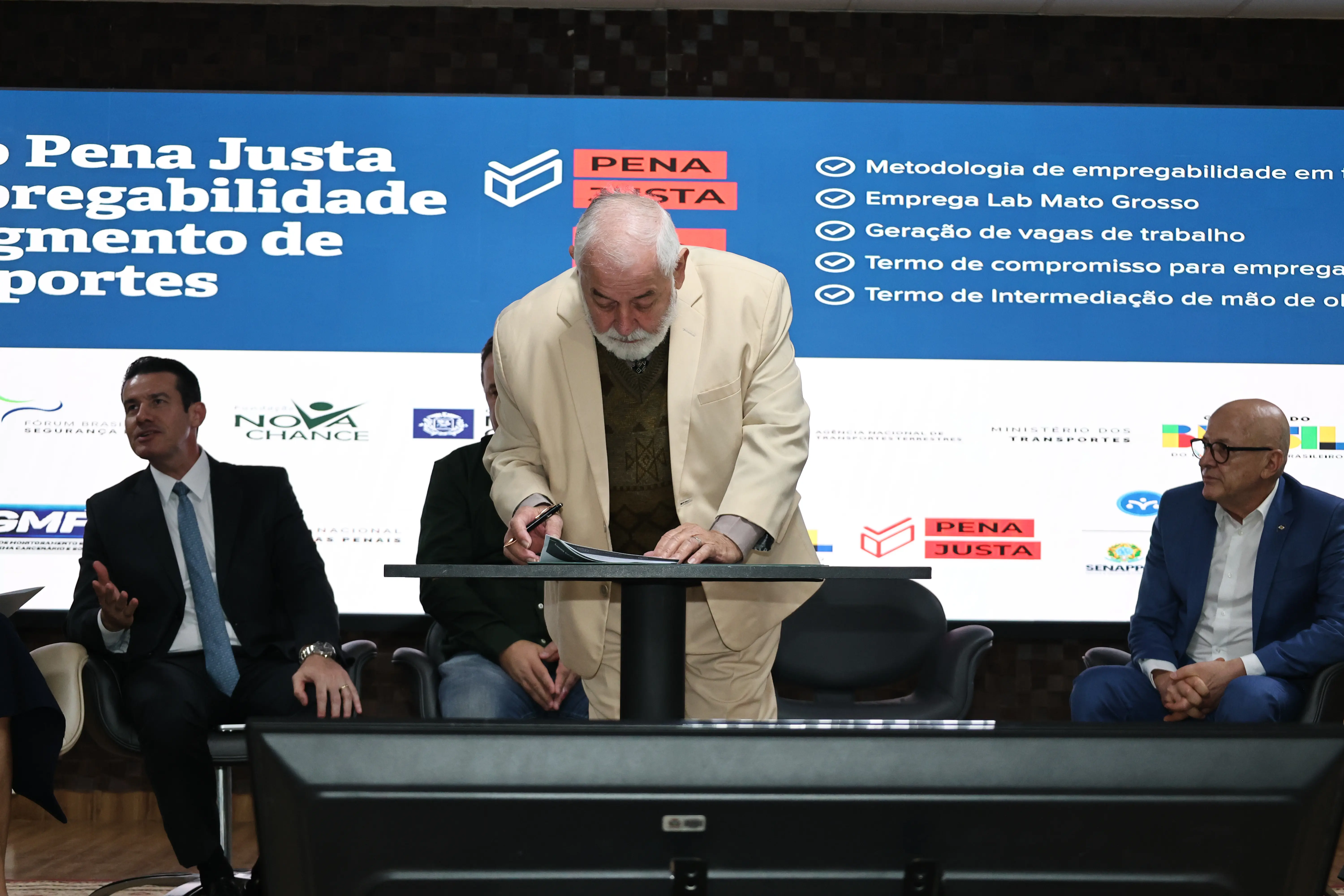 Homem branco, de terno bege inclinado sobre uma mesa, com uma caneta para assinatura de documentos. Ao fundo, telão exibe o texto 