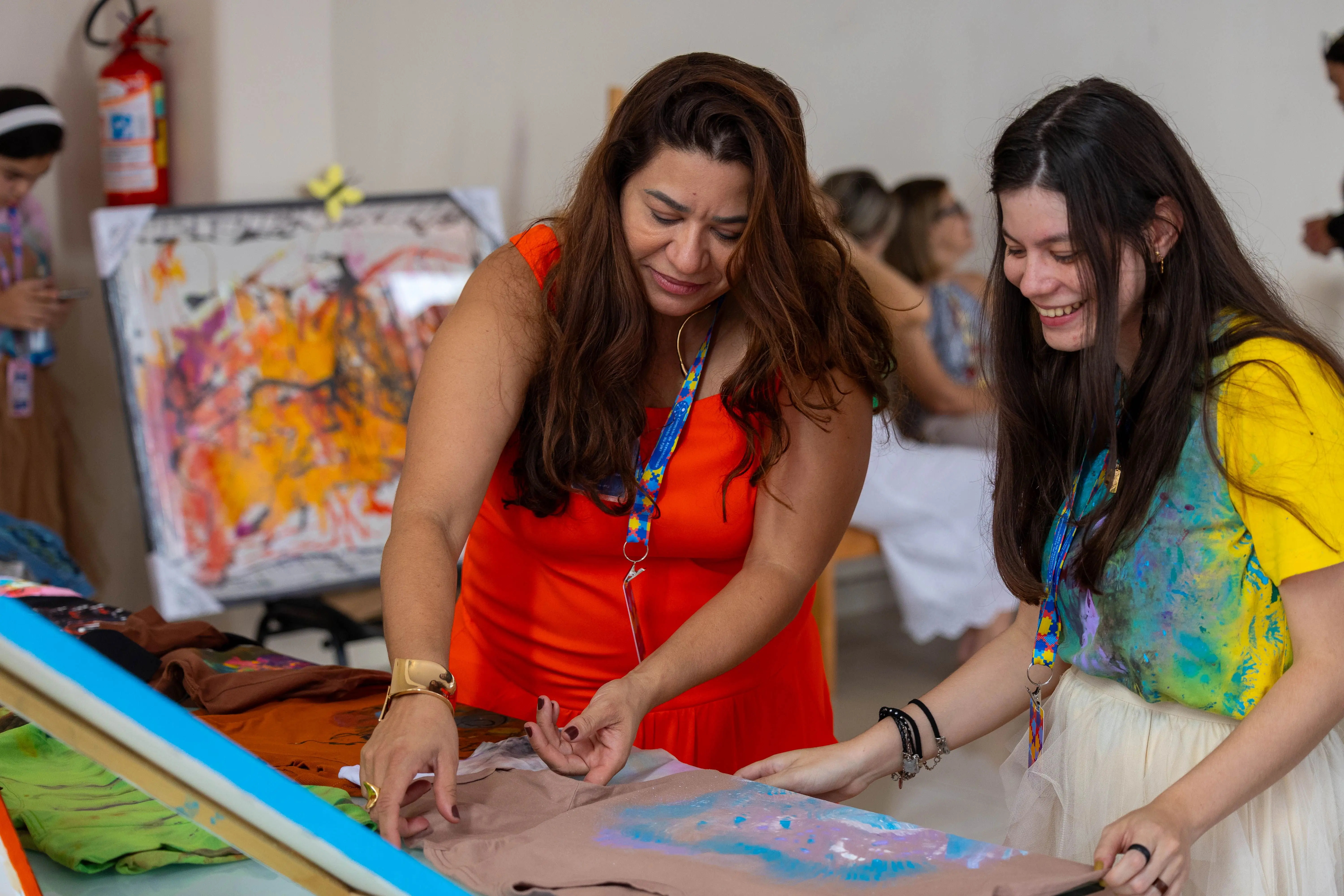Duas mulheres analisam camiseta com pintura artística sobre mesa. Cena mostra atividade criativa e interação, com ambiente leve e colaborativo.