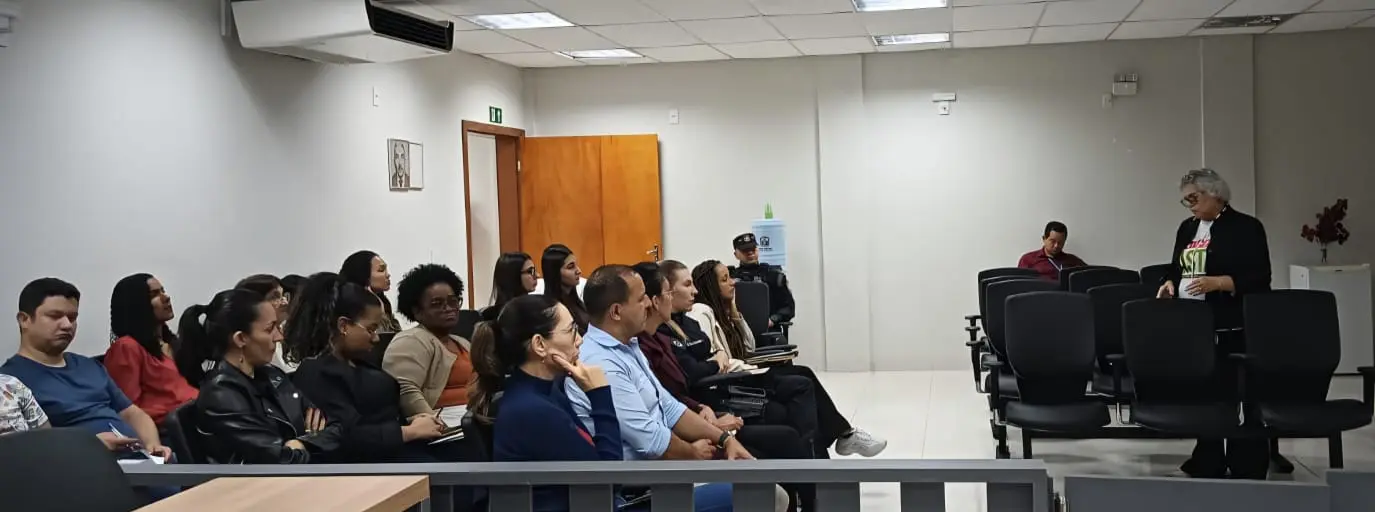 Participantes sentados em auditório assistem à apresentação de uma mulher de cabelos curtos e camiseta branca, de pé à direita. Ao fundo, uma porta de madeira e um bebedouro.