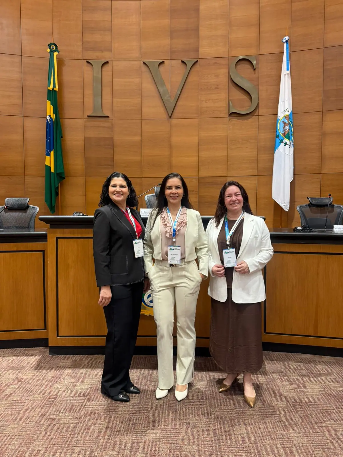 Três mulheres sorriem lado a lado em um plenário com painel de madeira ao fundo, onde se leem as letras 