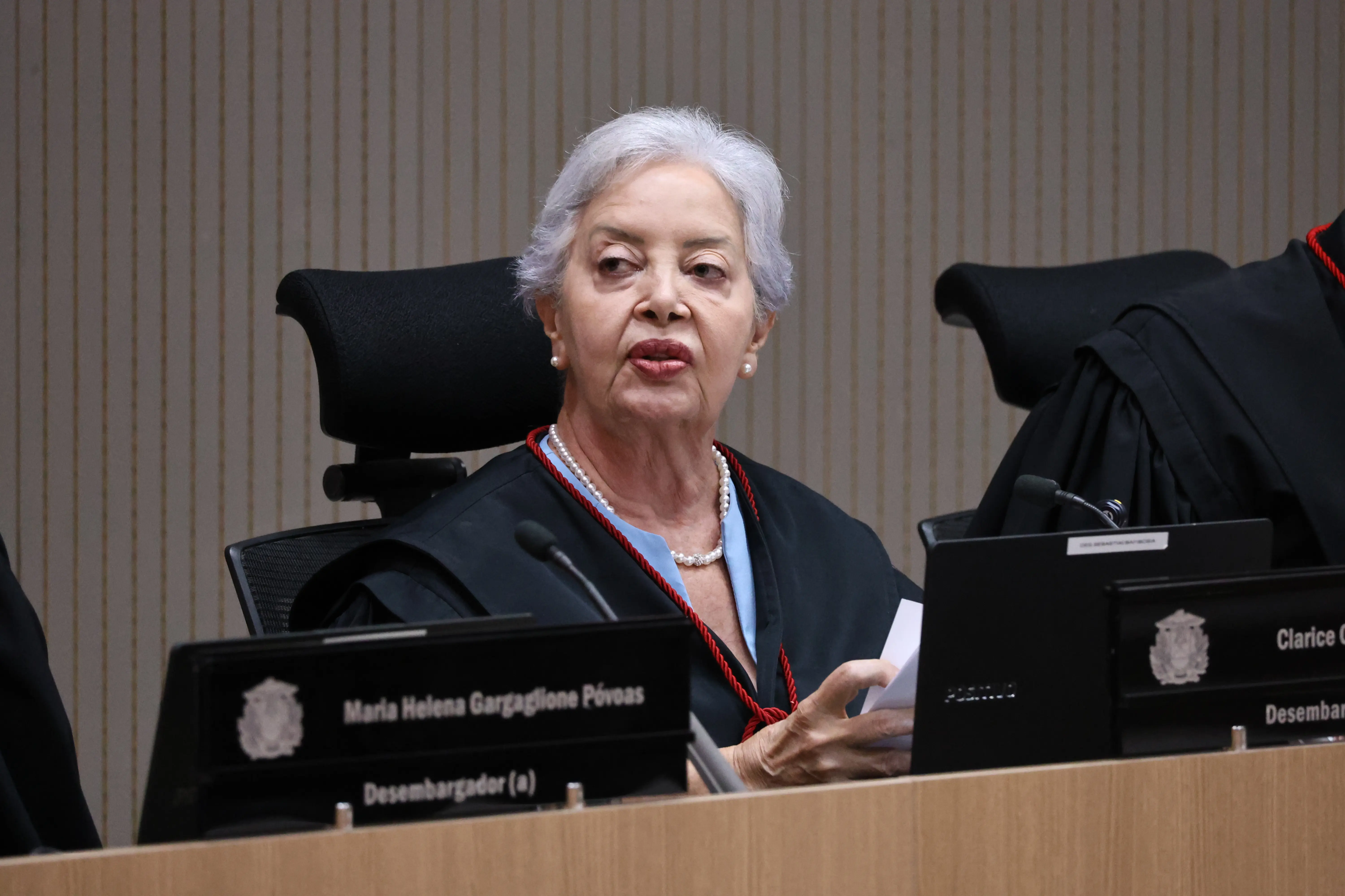 Mulher idosa de cabelos brancos curtos e toga preta sentada no plenário. À sua frente, placas de identificação e um notebook. Ela segura papéis e olha atentamente para o lado.