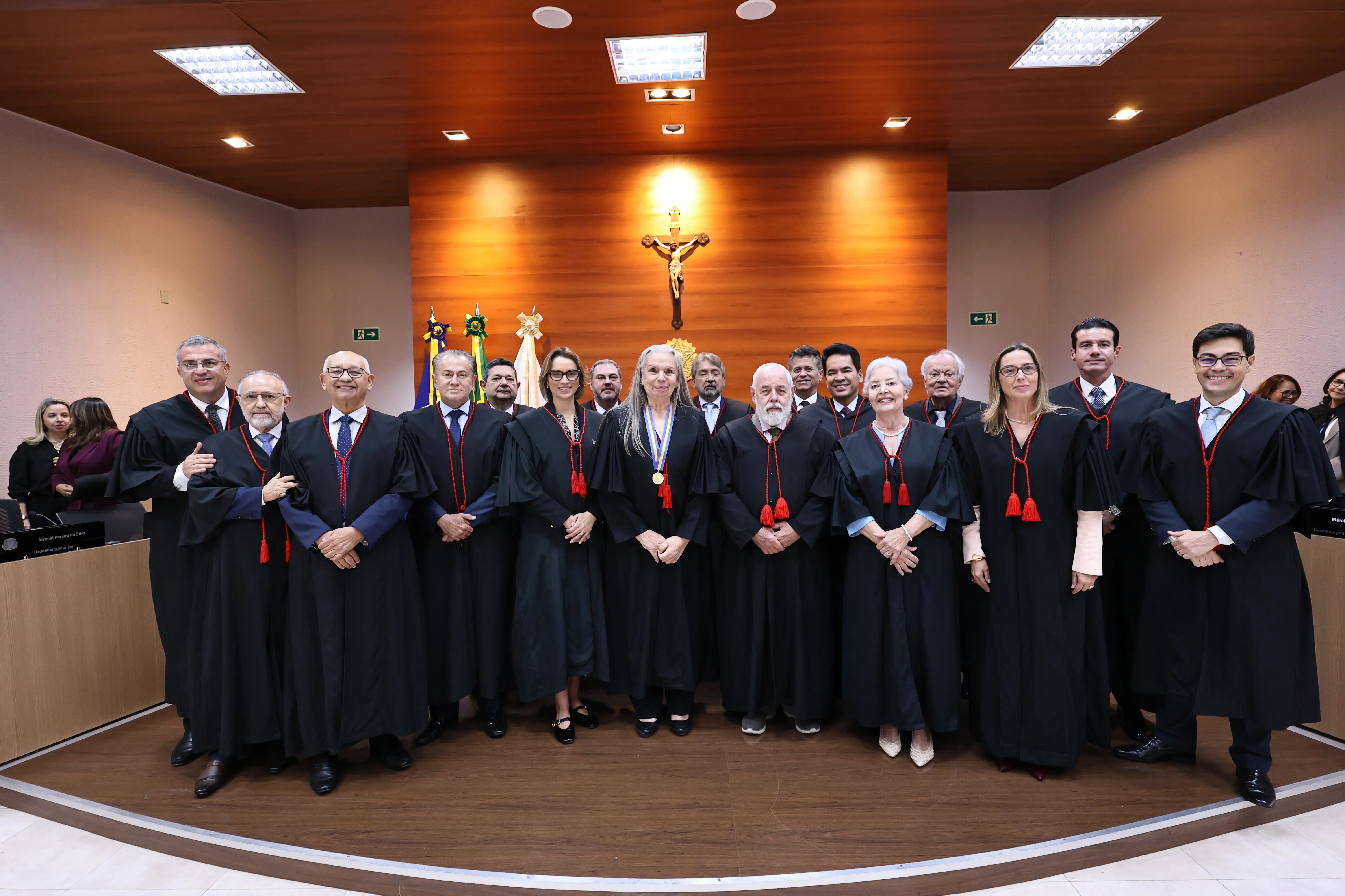 Grande grupo de magistrados de toga posa sorrindo lado a lado no plenário de um tribunal. Ao fundo, há um painel de madeira com um crucifixo fixado no centro e bandeiras oficiais ao lado.