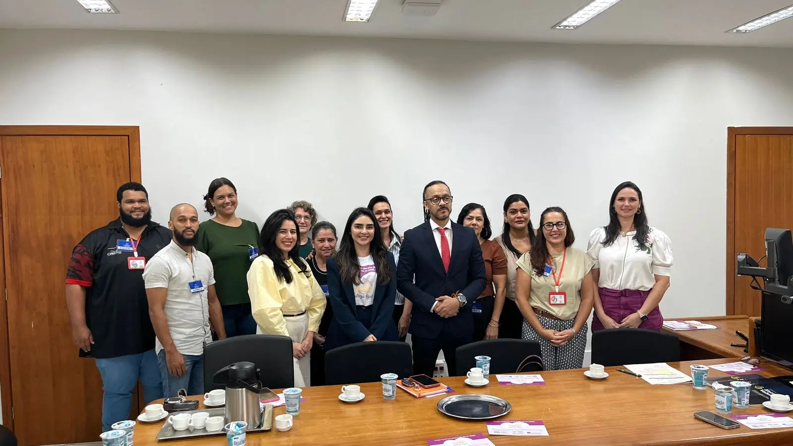 Foto posada de um grupo de 12 pessoas reunidas em uma sala de reuniões do Fórum de Juara. Os participantes estão posicionados atrás de uma mesa de madeira. Entre os presentes há homens e mulheres, alguns utilizando crachás de identificação. A sala possui paredes claras, portas e móveis de madeira.