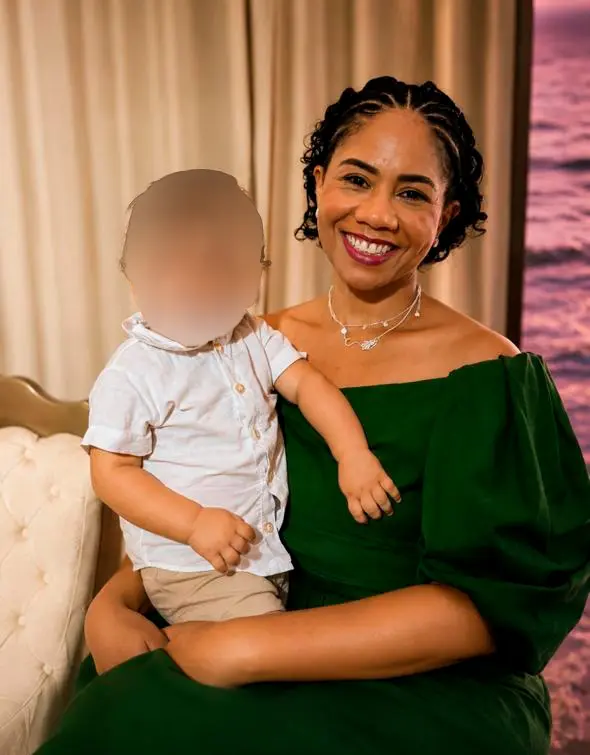 Mulher negra sorridente com cabelo preso e vestido verde escuro sentada, segurando no colo um bebê com o rosto borrado para preservação da imagem, de camisa social branca e bermuda bege. Ao fundo, uma cortina clara.