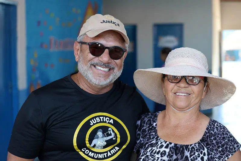 Foto horizontal colorida, que mostra o juiz coordenador da Justiça Comunitária, José Antonio Bezerra Filho, e a senhora Rita Maria Soares sorrindo para a foto, no pátio da escola onde ocorreu o mutirão Justiça em Ação. Ele usa boné bege, óculos de sol e camiseta preta da Justiça Comunitária.