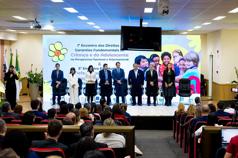 Foto horizontal colorida, em plano aberto, que mostra o auditório do Ministério Público repleto de pessoas sentadas. No palco, várias autoridades em pé. Atrás deles, há um enorme telão com a logomarca do evento: 1º Encontro dos Direitos e Garantias Fundamentais da Criança e do Adolescente.