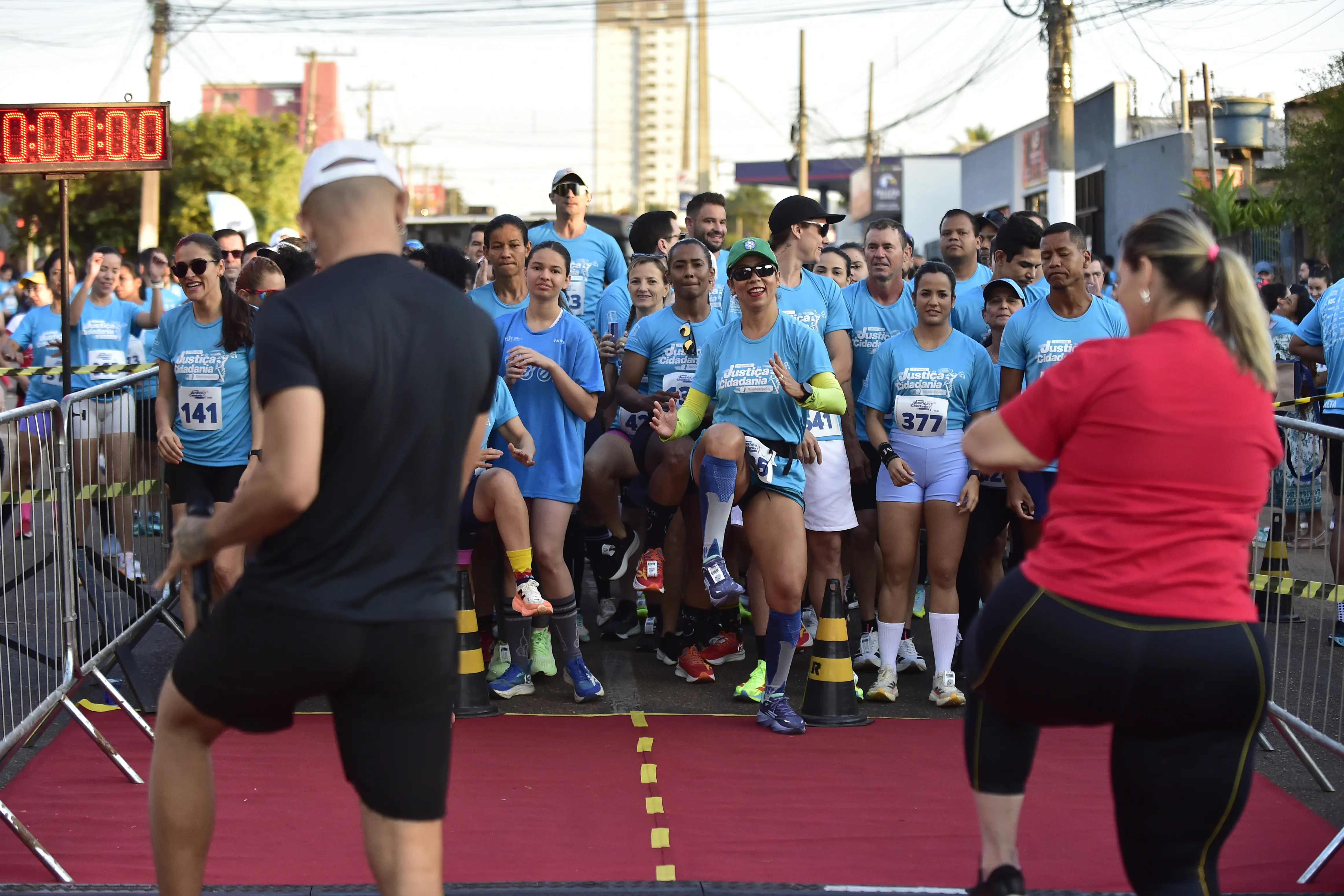 Grupo de corredores com camisas azuis se alonga na linha de largada de uma corrida de rua. Dois instrutores de costas, em cima de um tapete vermelho, orientam os exercícios de aquecimento.