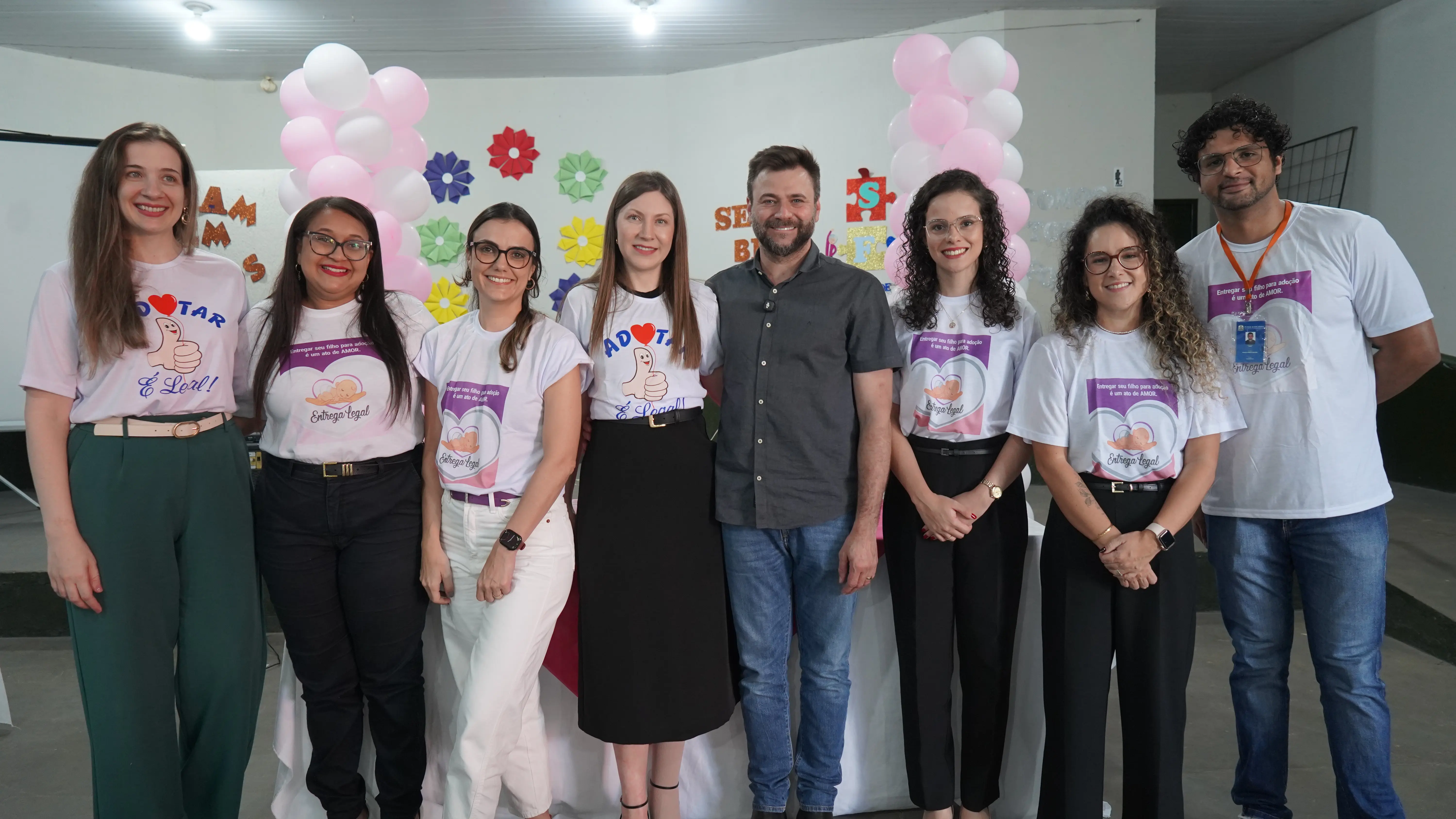 Grupo de oito pessoas sorrindo lado a lado em um evento sobre adoção. Vestem camisetas brancas da campanha 