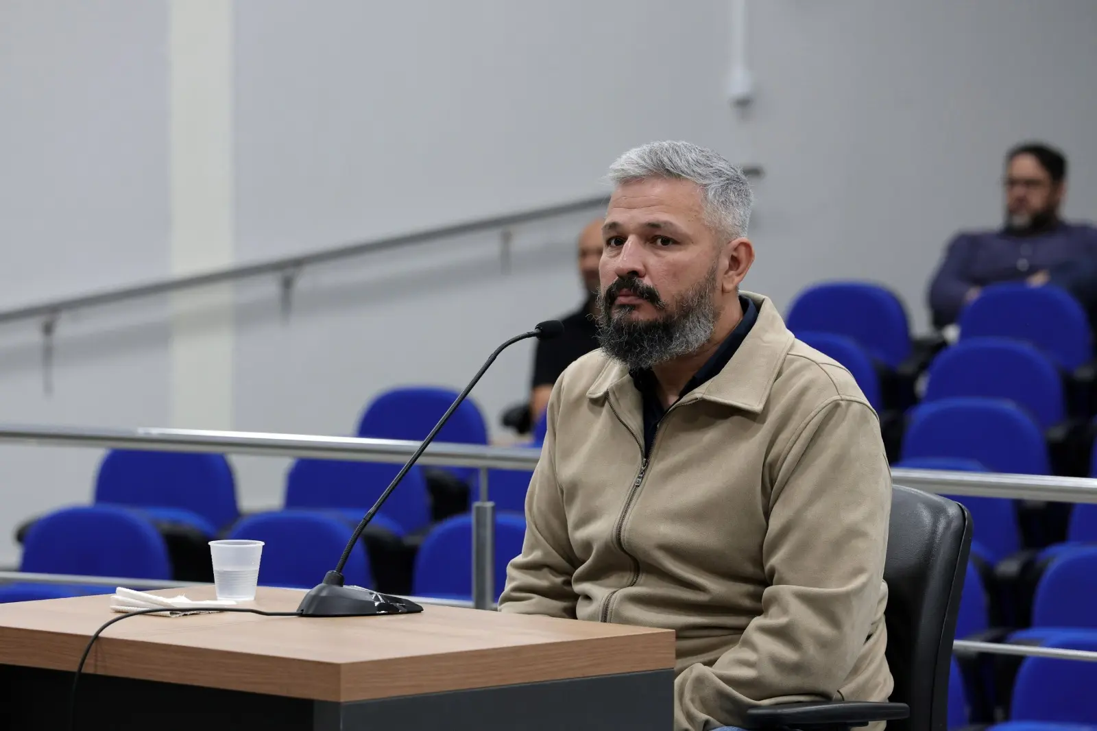 Foto horizontal colorida em plano médio que mostra o policial civil Mário Wilson sentado no banco dos réus no Tribunal do Júri. Ele é um homem pardo, alto, de cabelos e barba grisalhos, usando jaqueta bege.