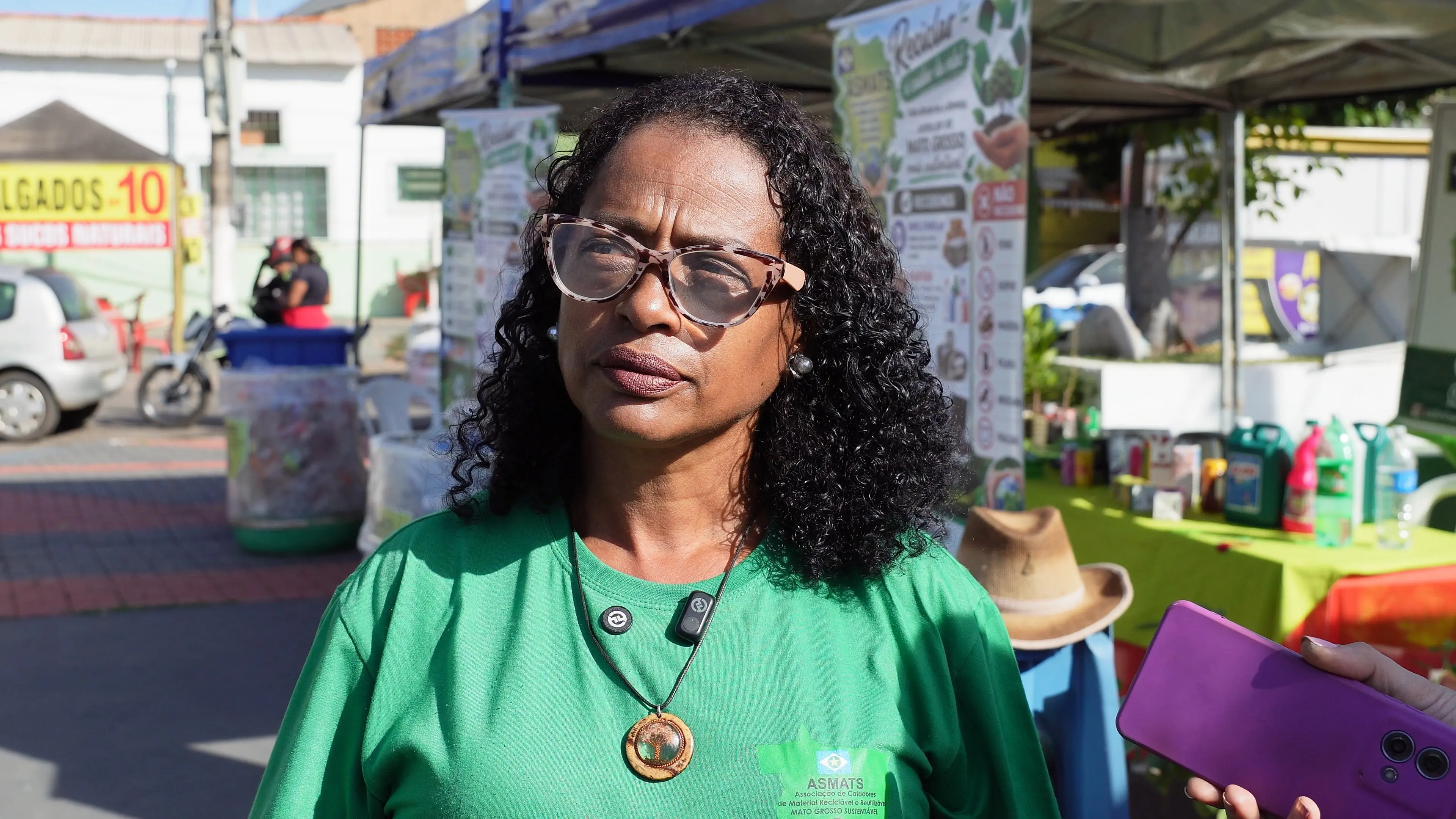 Mulher de blusa verde e óculos concede entrevista em área destinada à coleta de recicláveis. Ao fundo, cartazes explicam os tipos de materiais aceitos para reciclagem.